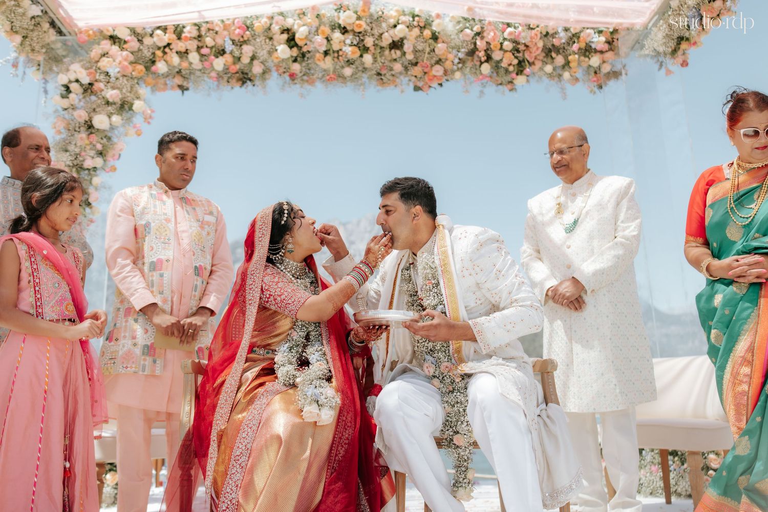 Indian bride feeds groom during vibrant Hindu wedding ceremony under floral mandap with family gathered.