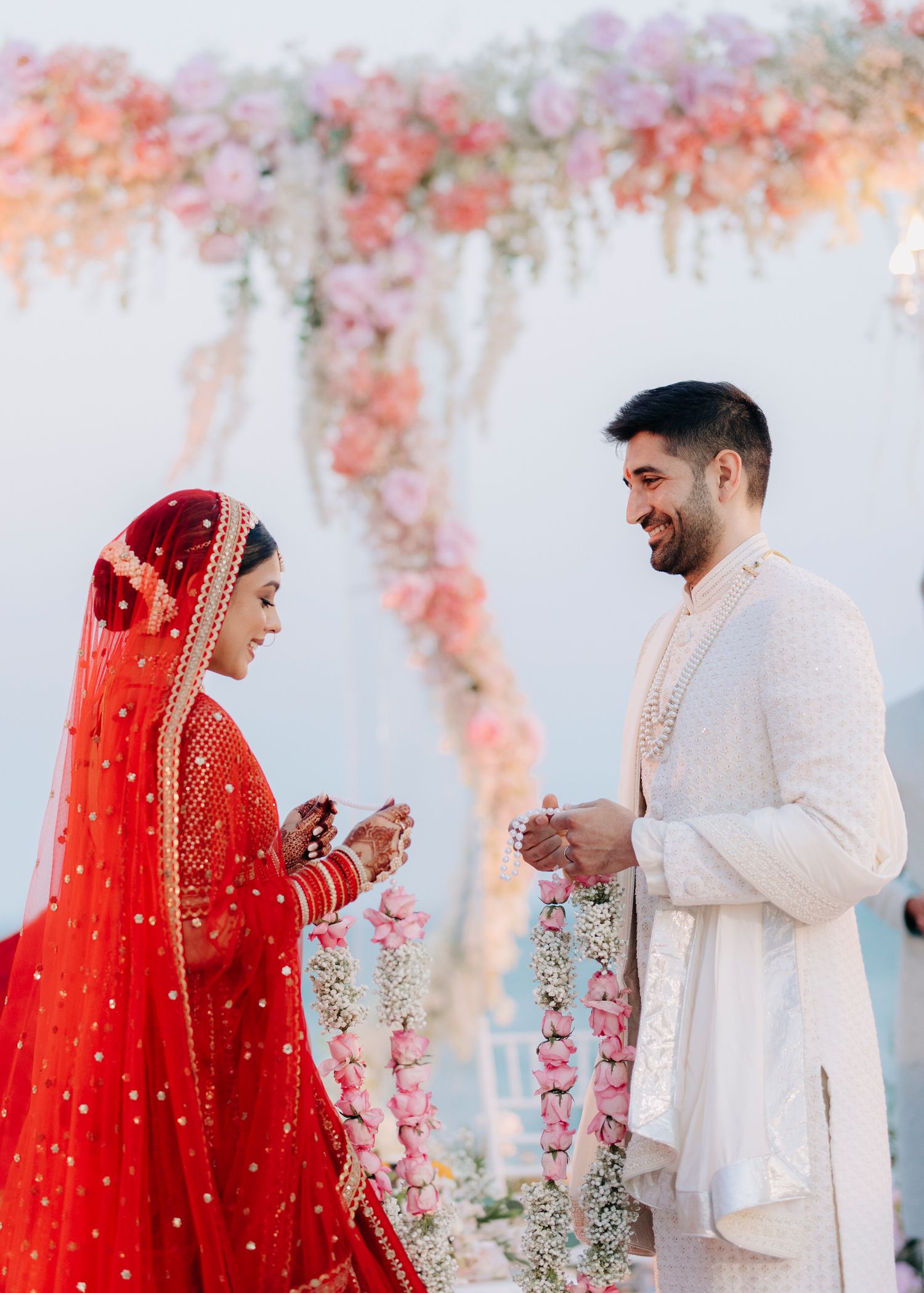 A couple in traditional Indian wedding attire exchanges rings under a floral arch with coral and pink blooms.
