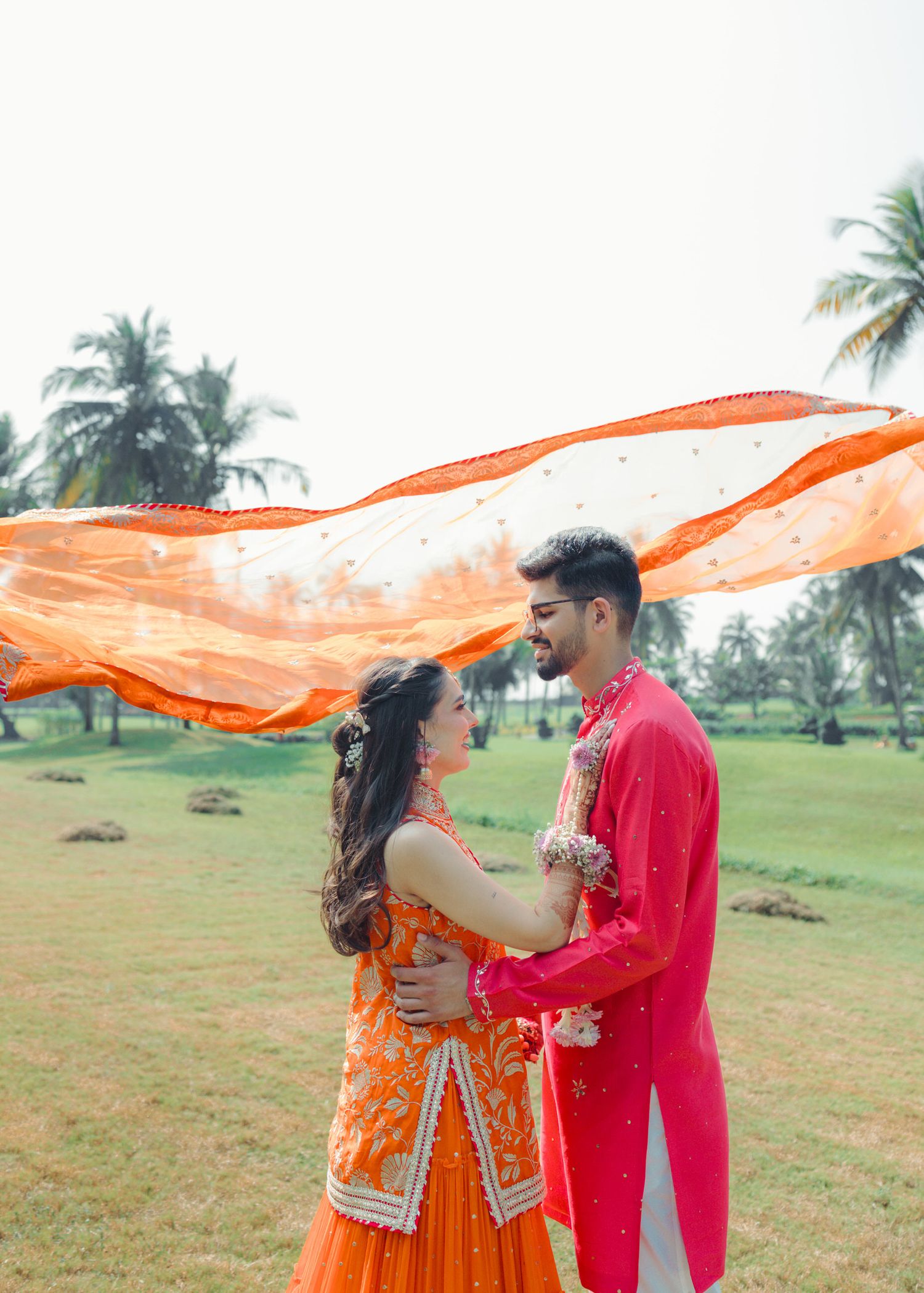 A couple in orange and red traditional attire poses outdoors with flowing fabric against a palm tree backdrop.