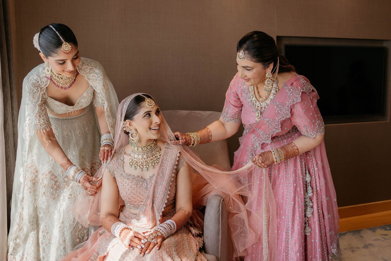 Three women wearing traditional South Asian wedding attire help prepare a bride in an elegant pink and white lehenga.