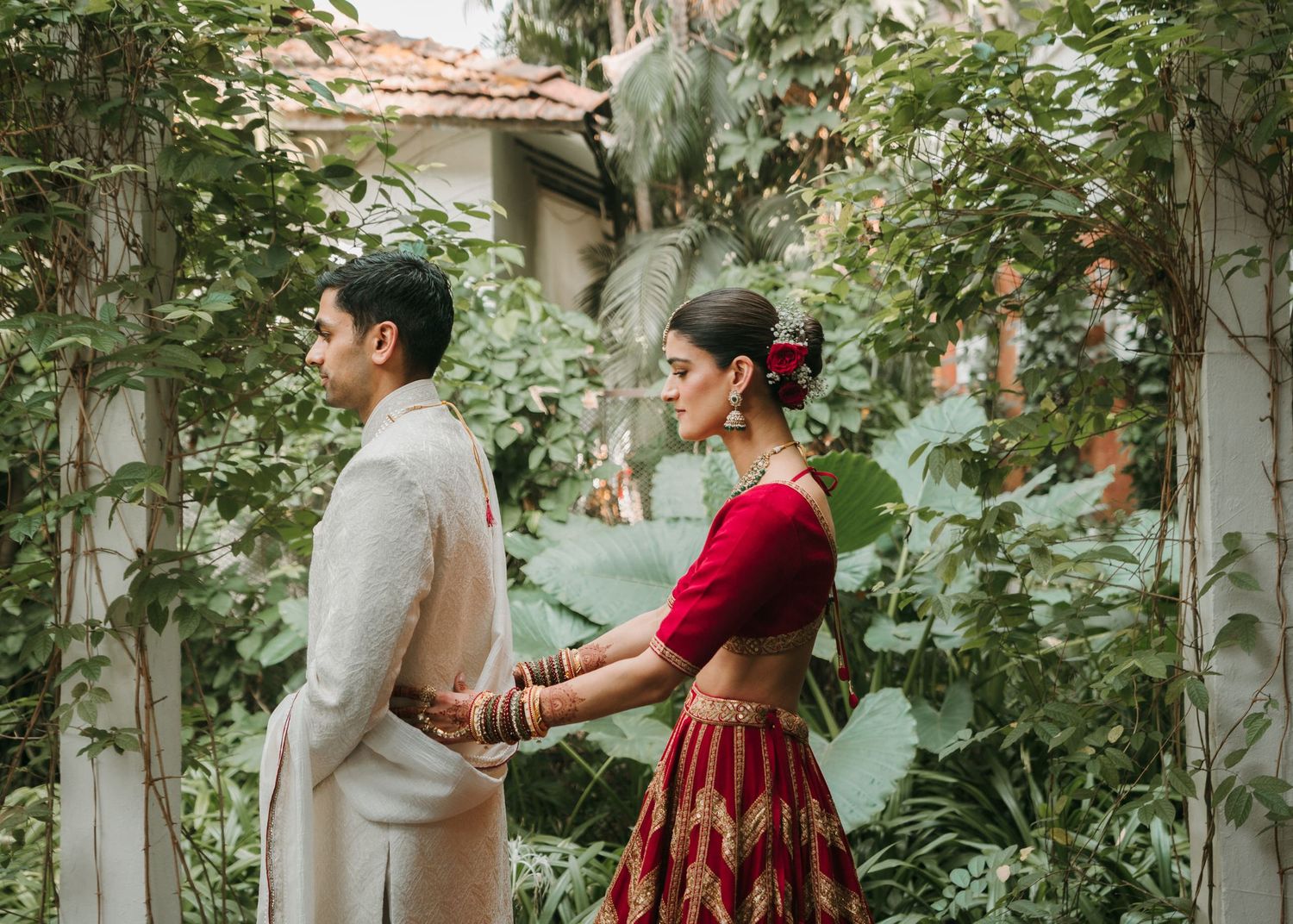 A couple in traditional Indian attire standing back to back in a lush garden setting with a rustic wall.