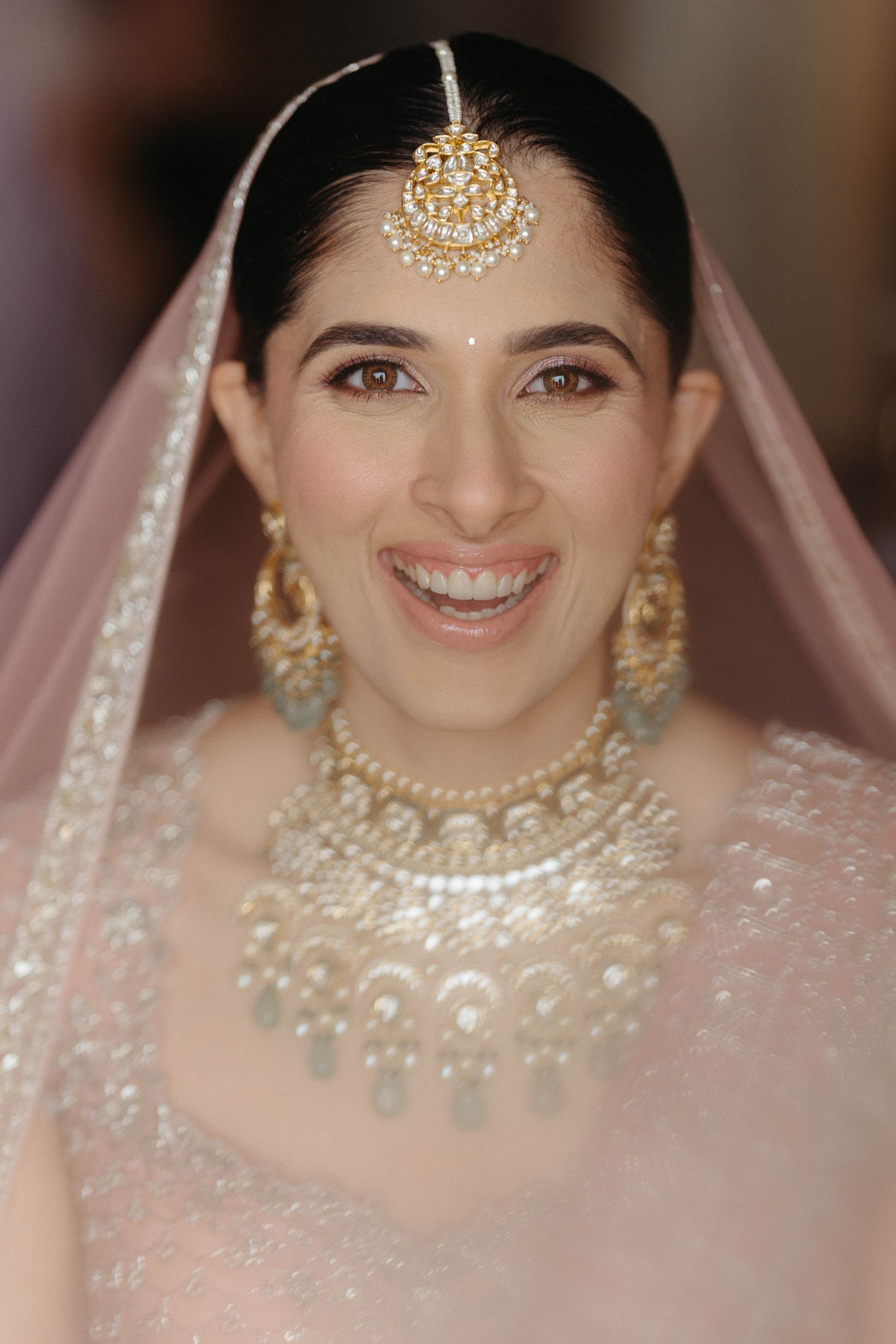 Indian bride wearing traditional gold bridal jewelry with pearls and a blush pink veil smiling at camera.