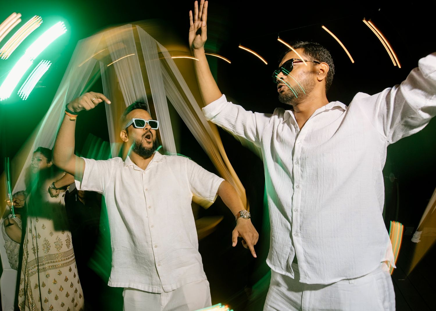 People dancing and celebrating in white attire under green lighting at a wedding reception.