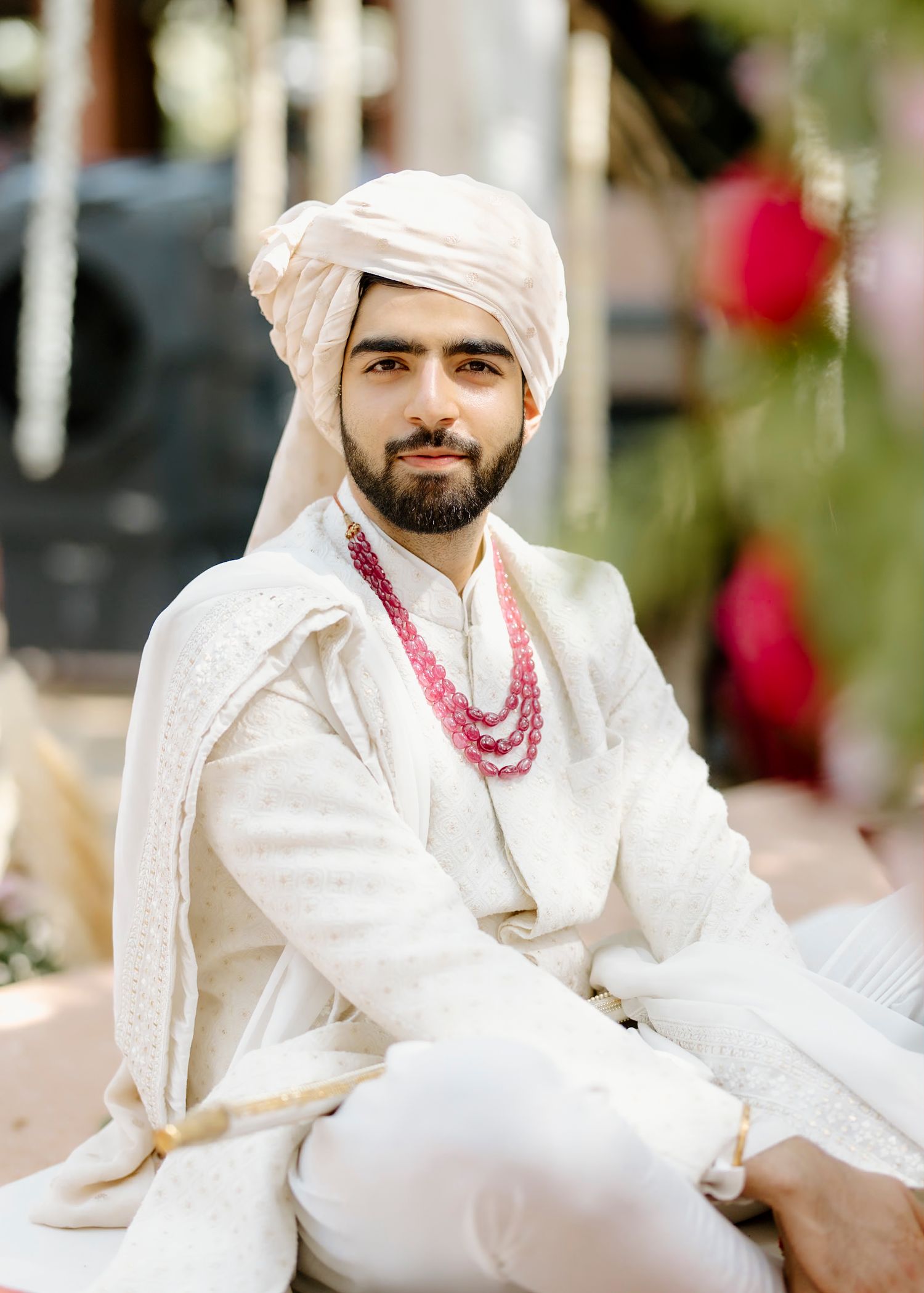 Traditional groom in white attire with pink accessories seated during wedding ceremony.