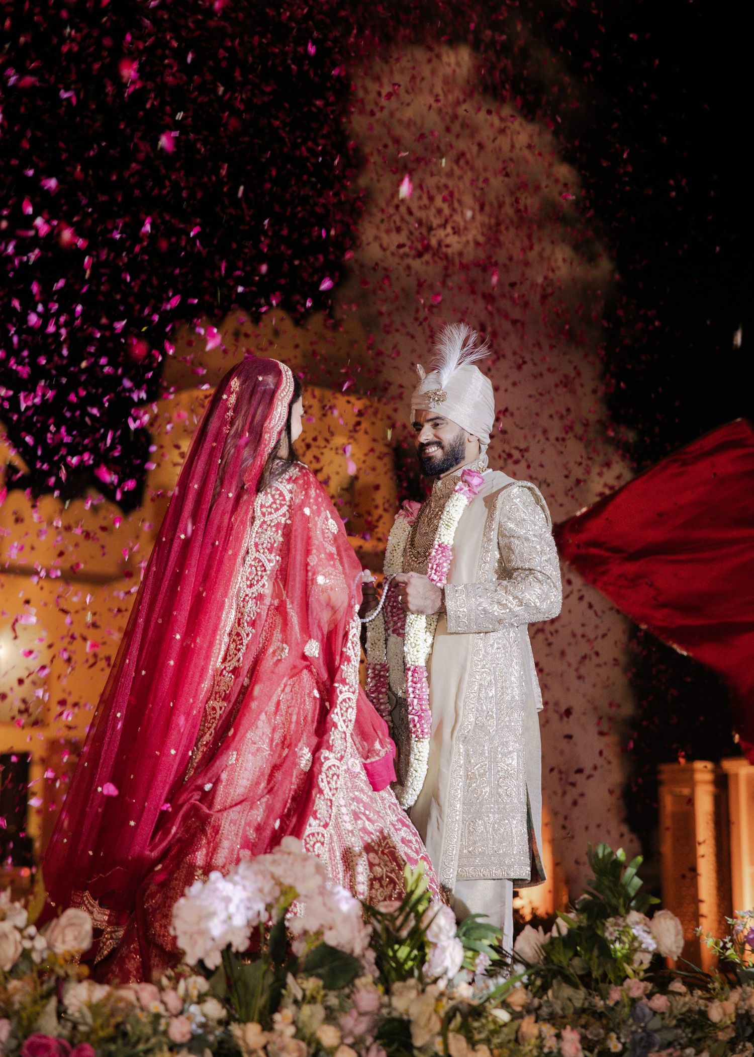 Indian bride in red lehenga and groom in white sherwani exchange garlands amid falling rose petals.