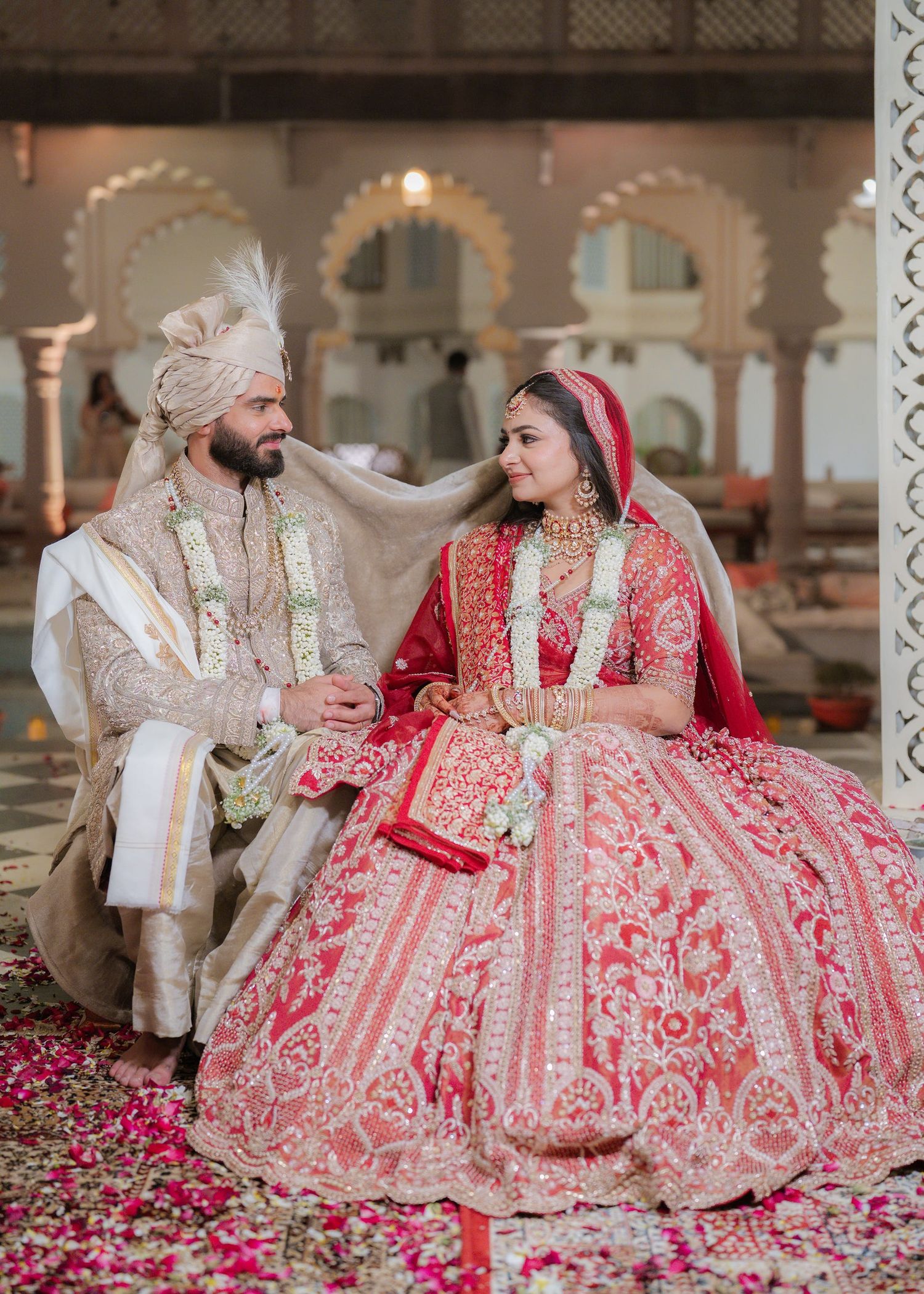 Indian bride in red lehenga and groom in ivory sherwani share a tender moment at traditional Hindu wedding ceremony.