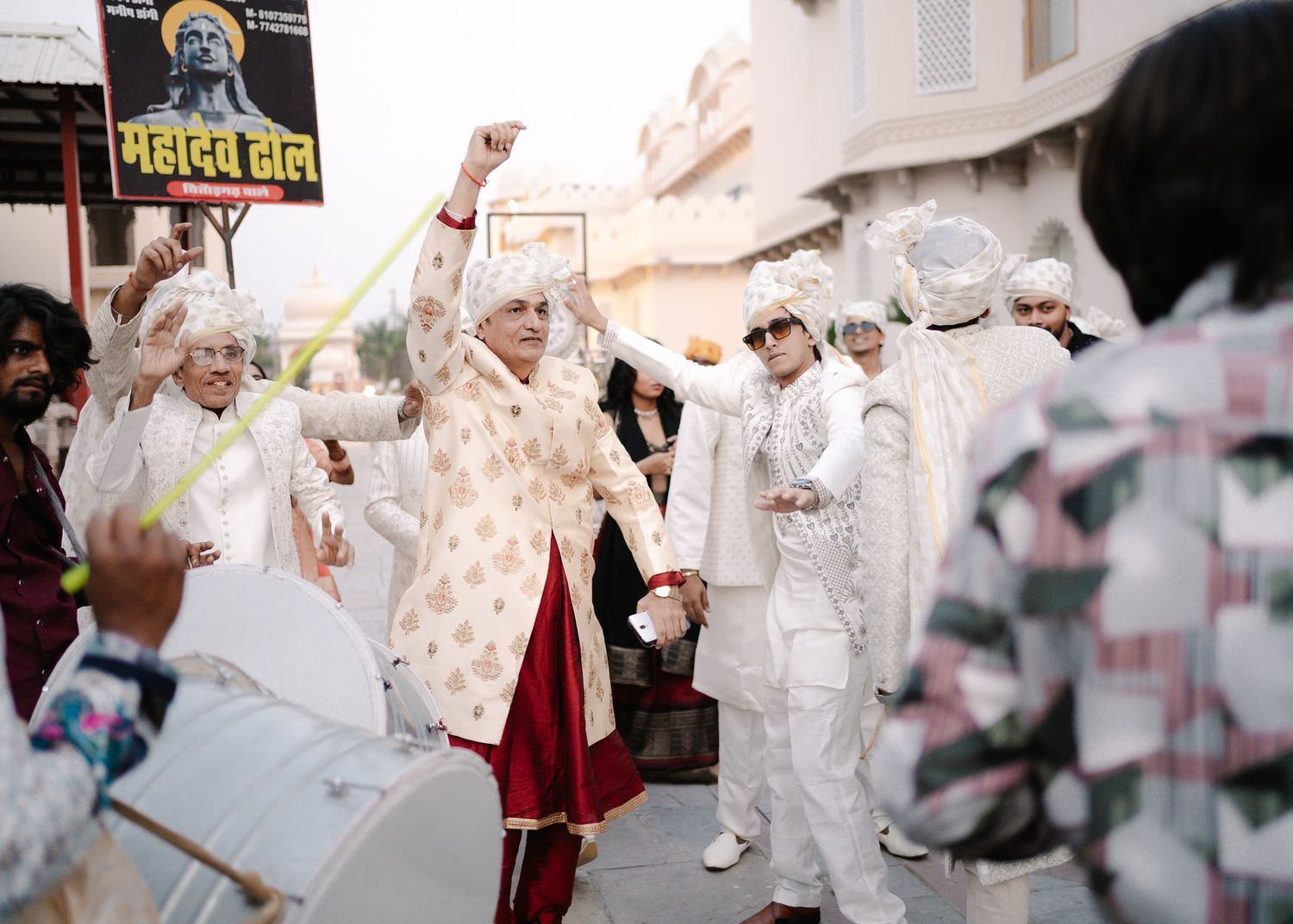 Guests in white outfits celebrate with music and dancing during a vibrant Indian wedding baraat procession.