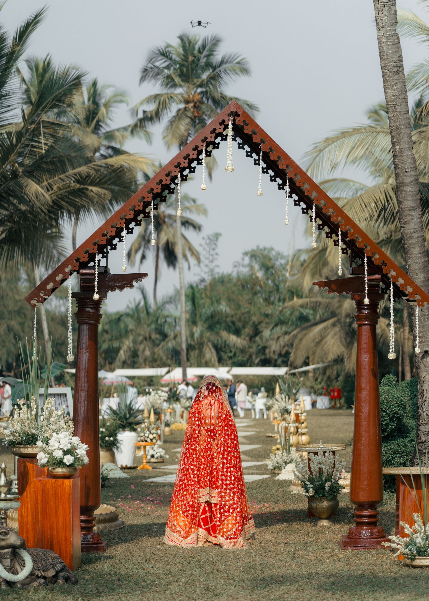 Sequence of wooden gazebos decorated with red and white fabric and hanging lights amid palm trees at a tropical venue.