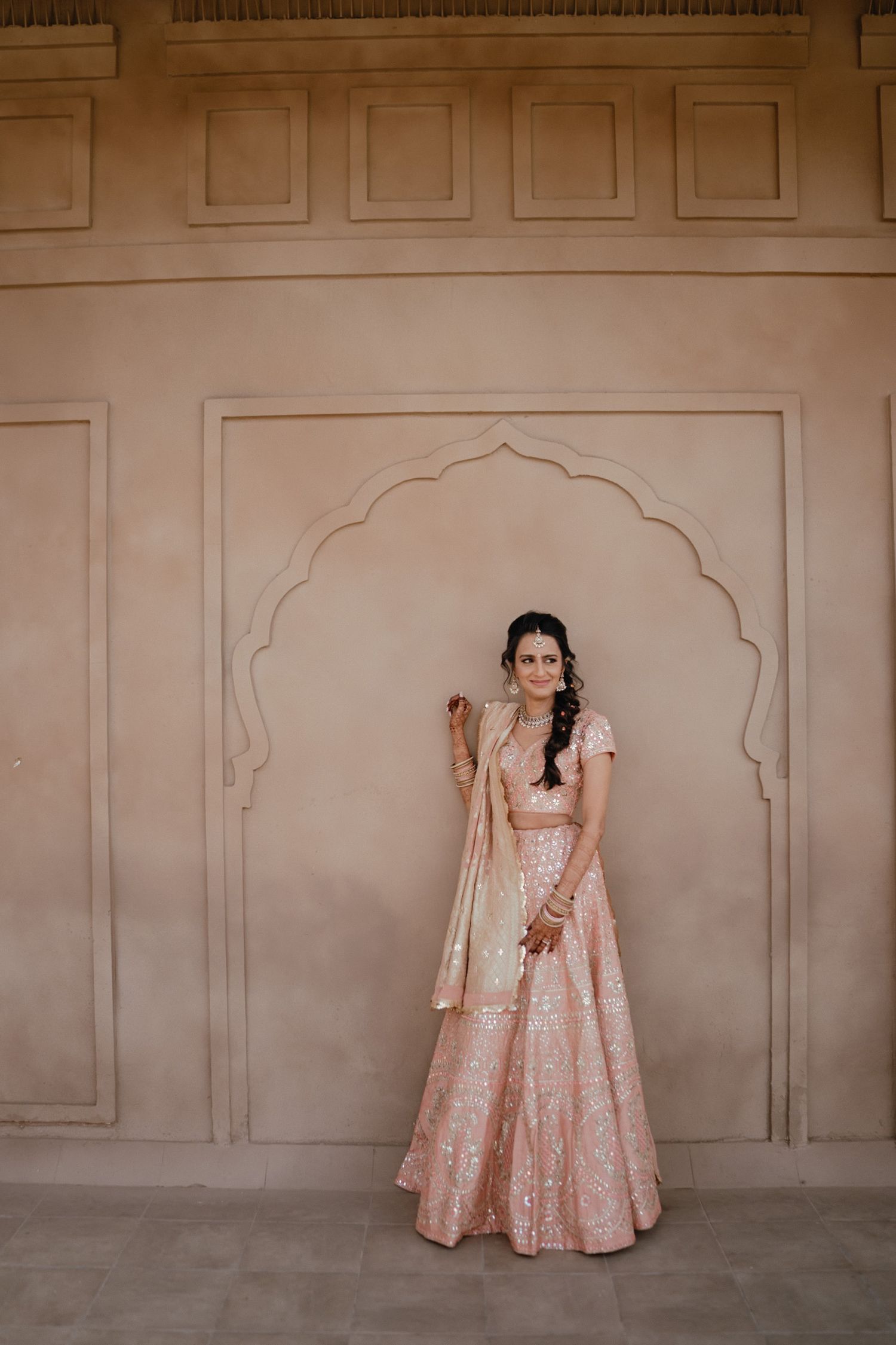 Traditional South Asian bridal attire featuring a pink embroidered lehenga against an ornate archway backdrop.