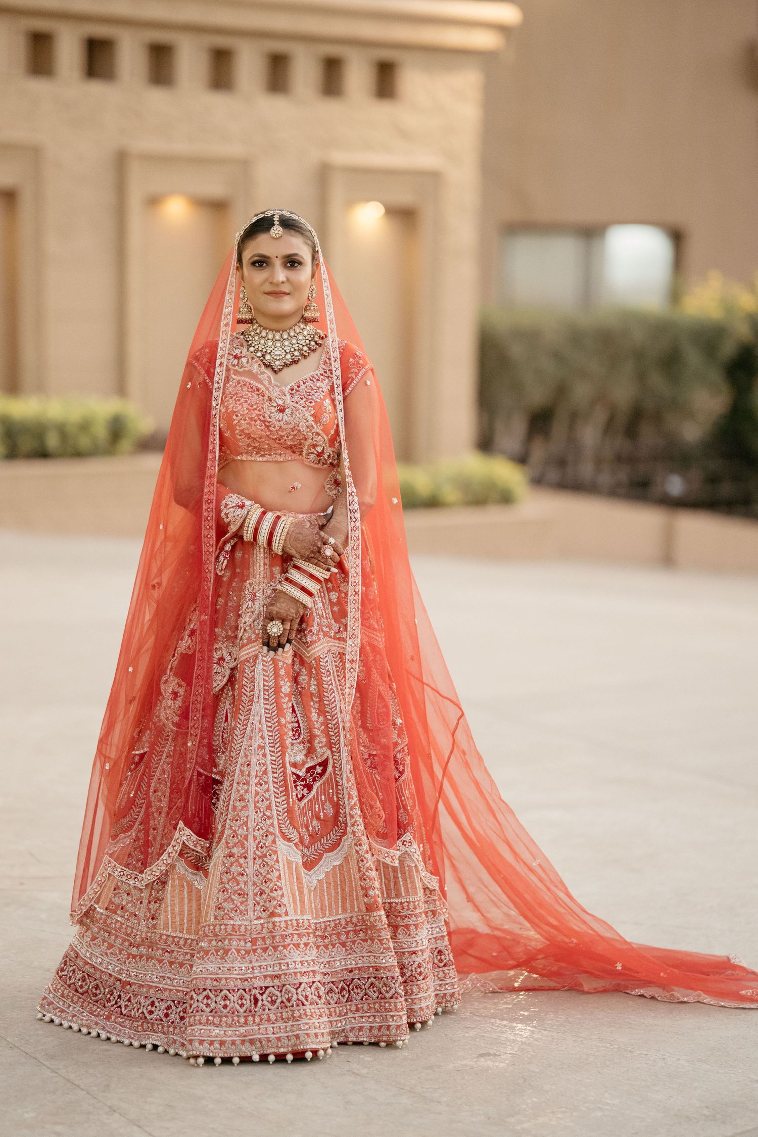 Beautiful bridal lehenga in coral red with intricate embroidery and matching sheer dupatta at wedding venue.