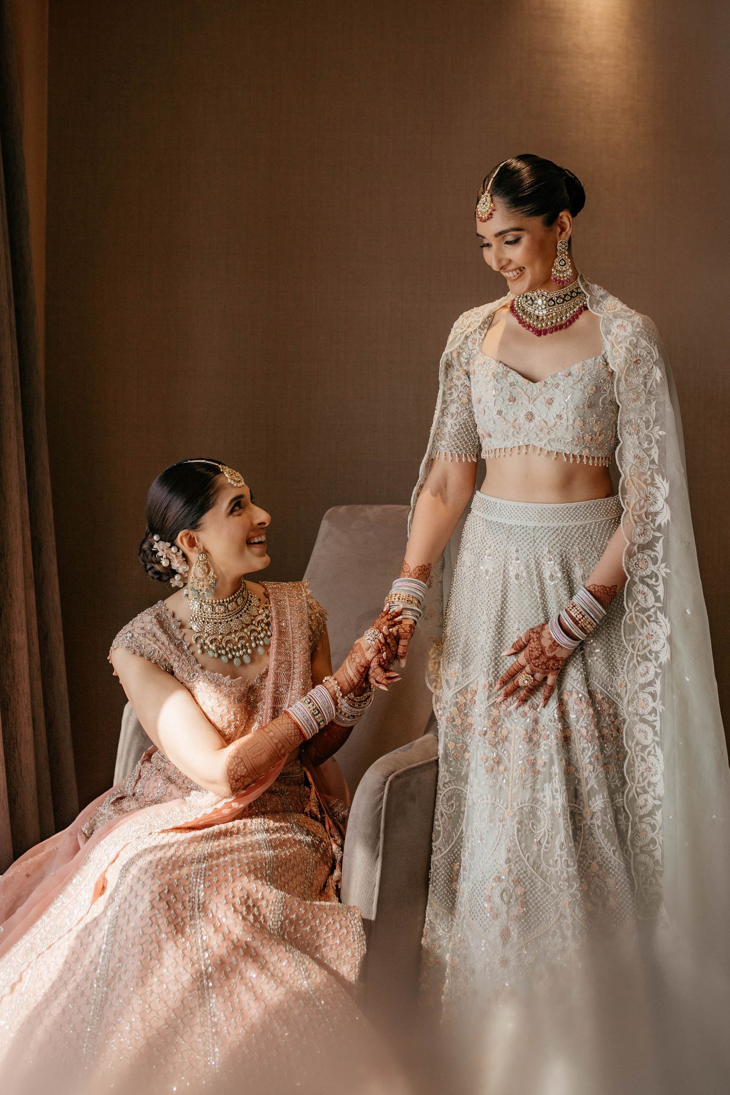 Two women in traditional Indian wedding attire share a candid moment in warm lighting.