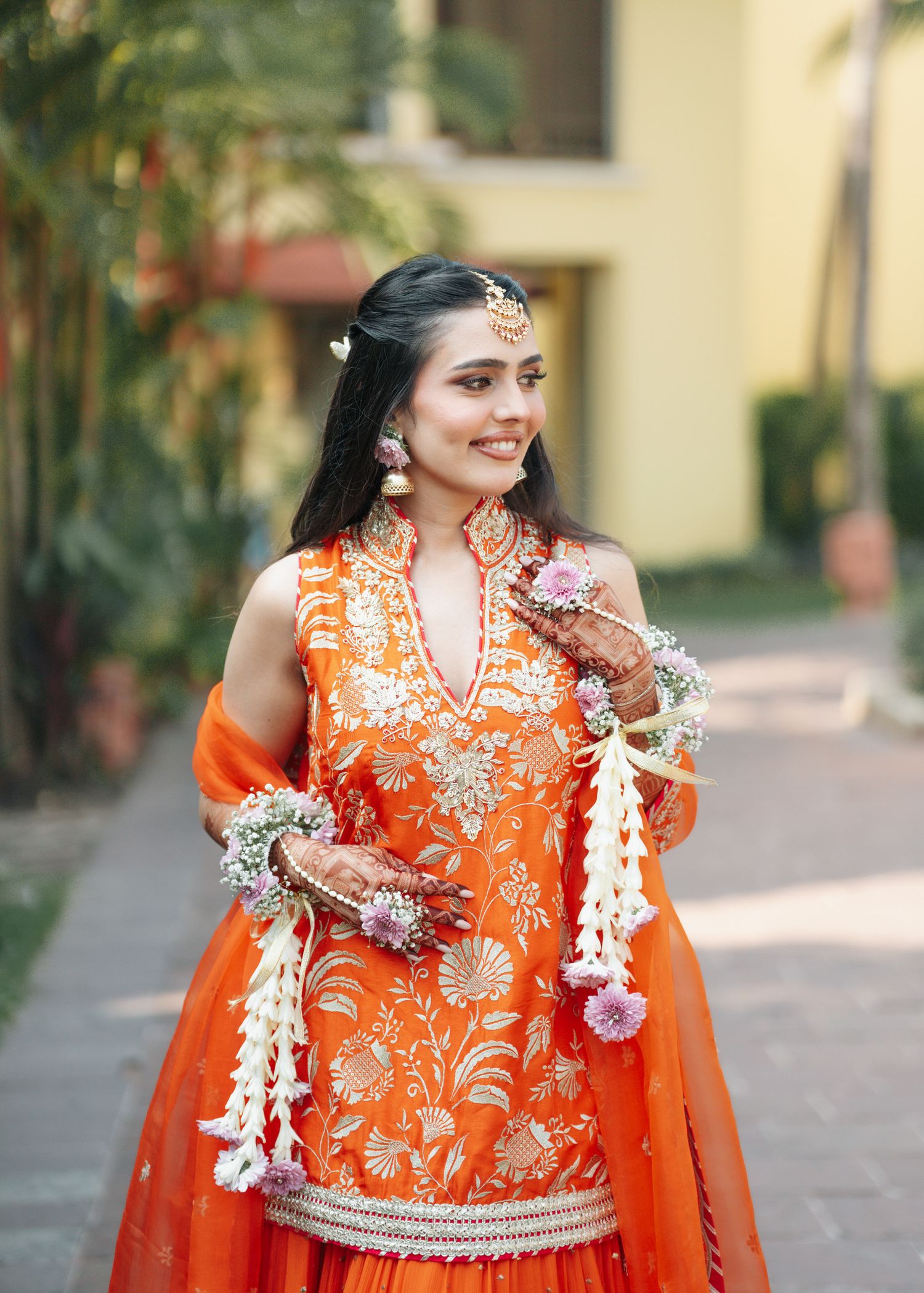 Traditional Indian bridal attire in bright orange with intricate embroidery and white floral garlands against yellow building.