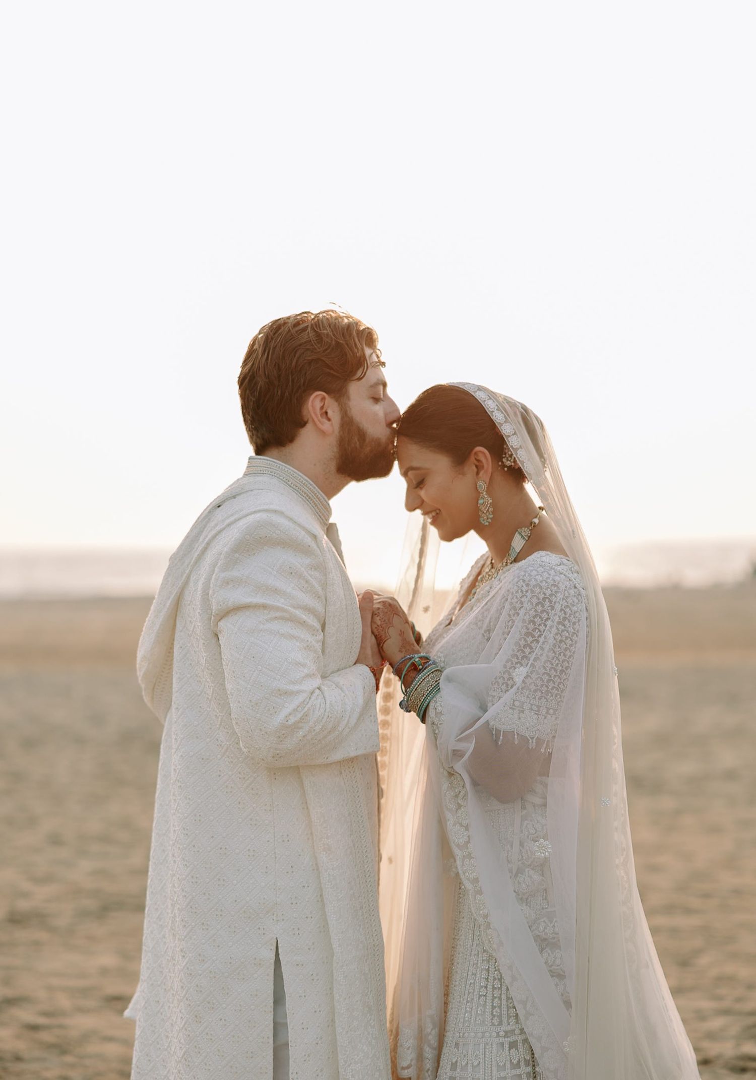 A couple in white wedding attire shares a tender kiss on a sandy beach at golden hour.