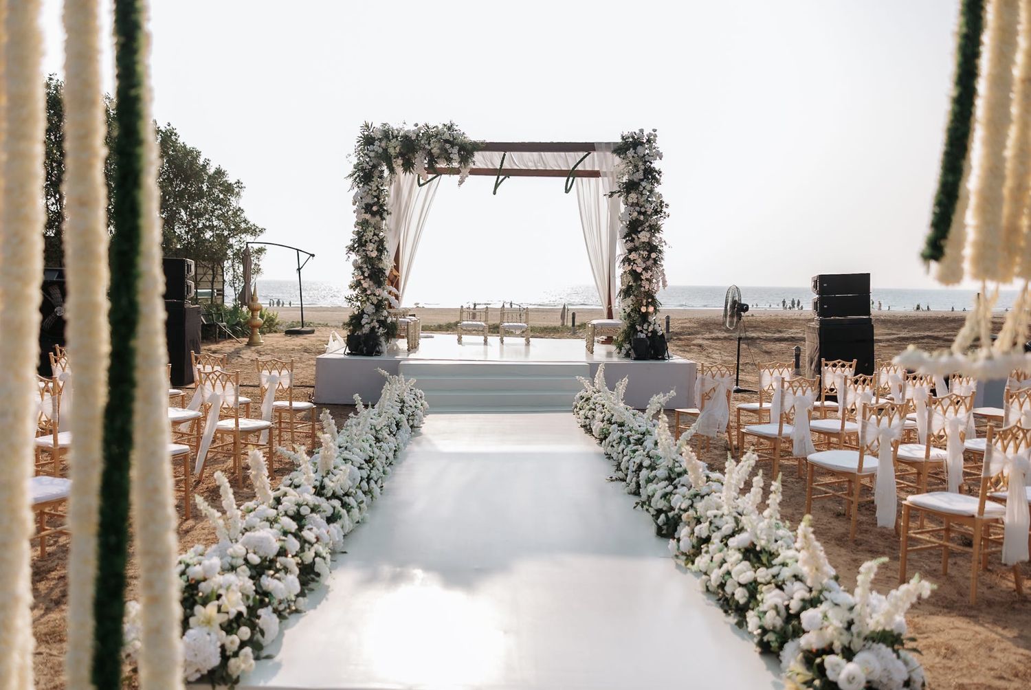 Elegant outdoor beach wedding ceremony aisle with white floral arrangements and a wooden floral arch.