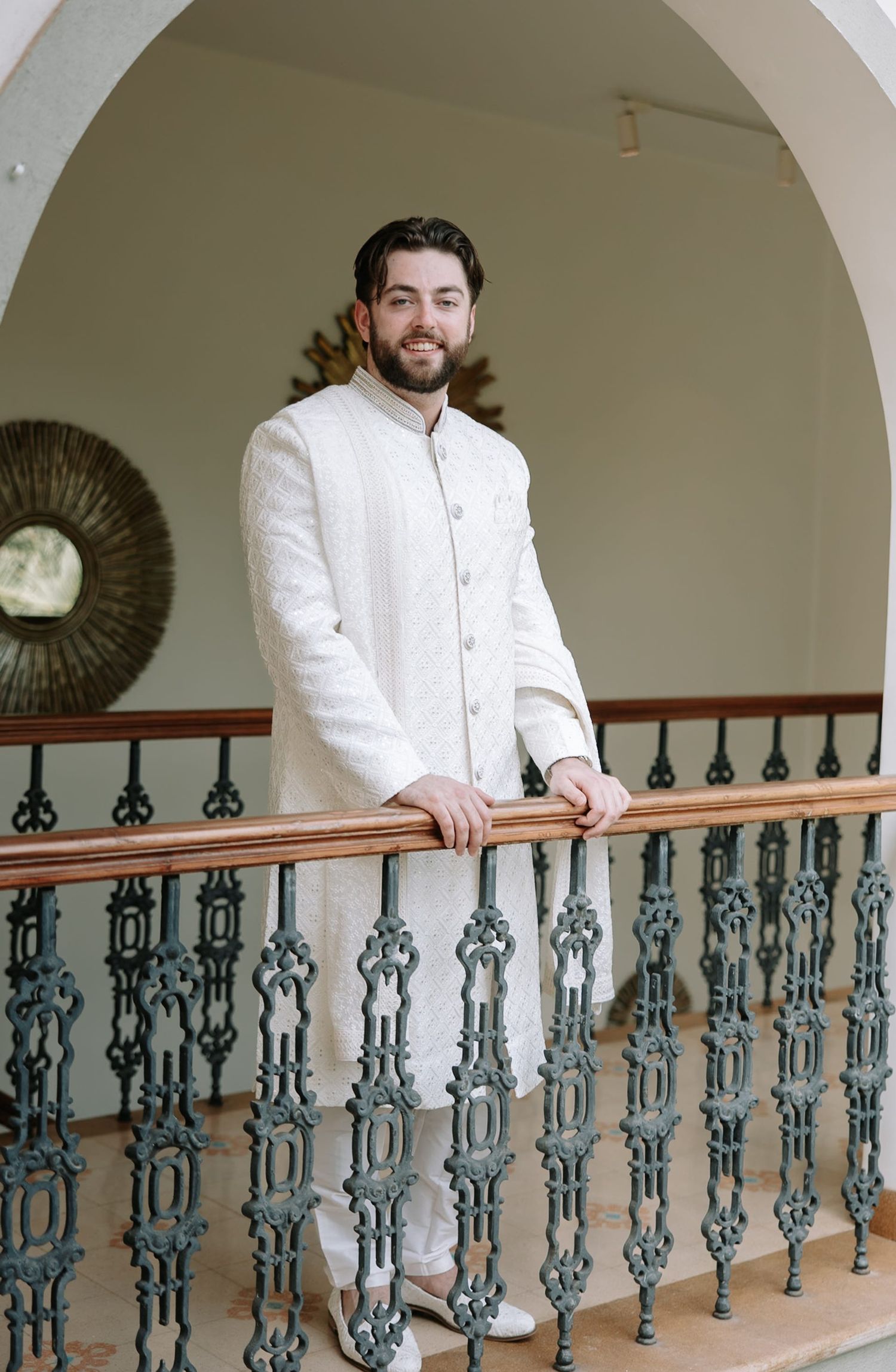 Man in white sherwani suit posing on ornate balcony with decorative iron railings at elegant venue.
