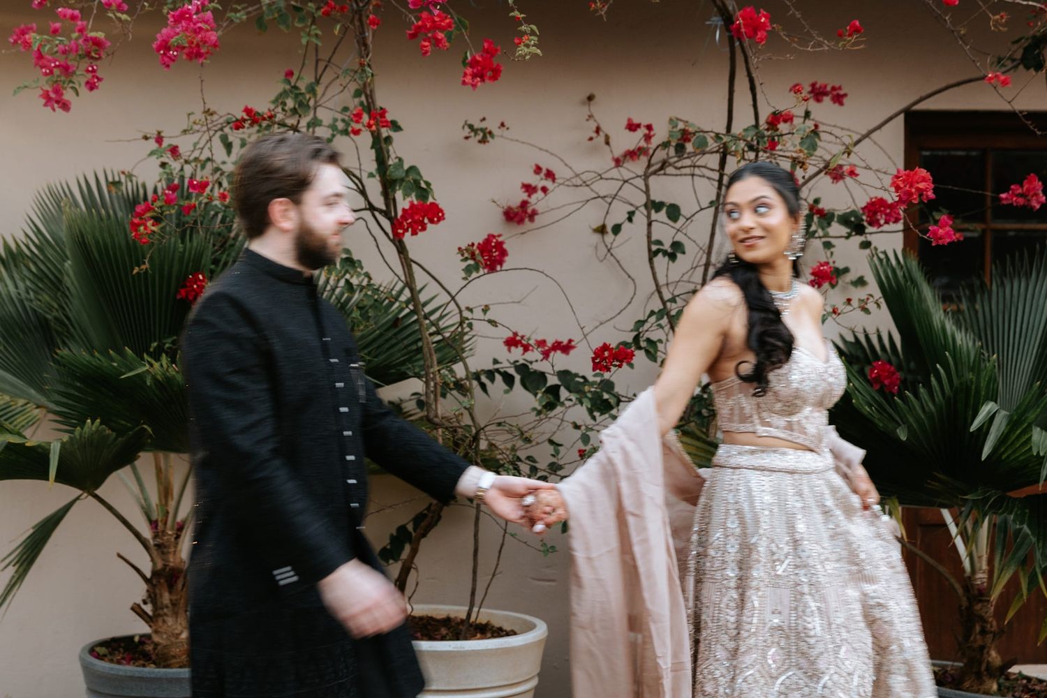 Couple in elegant attire holding hands outdoors, woman in beaded lehenga, man in black sherwani, red bougainvillea backdrop.
