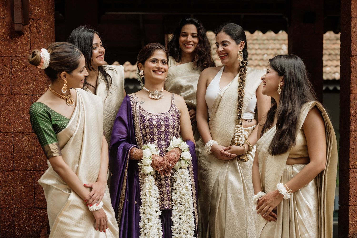 Bride in purple saree with jasmine garland surrounded by bridesmaids in cream sarees at South Indian wedding.