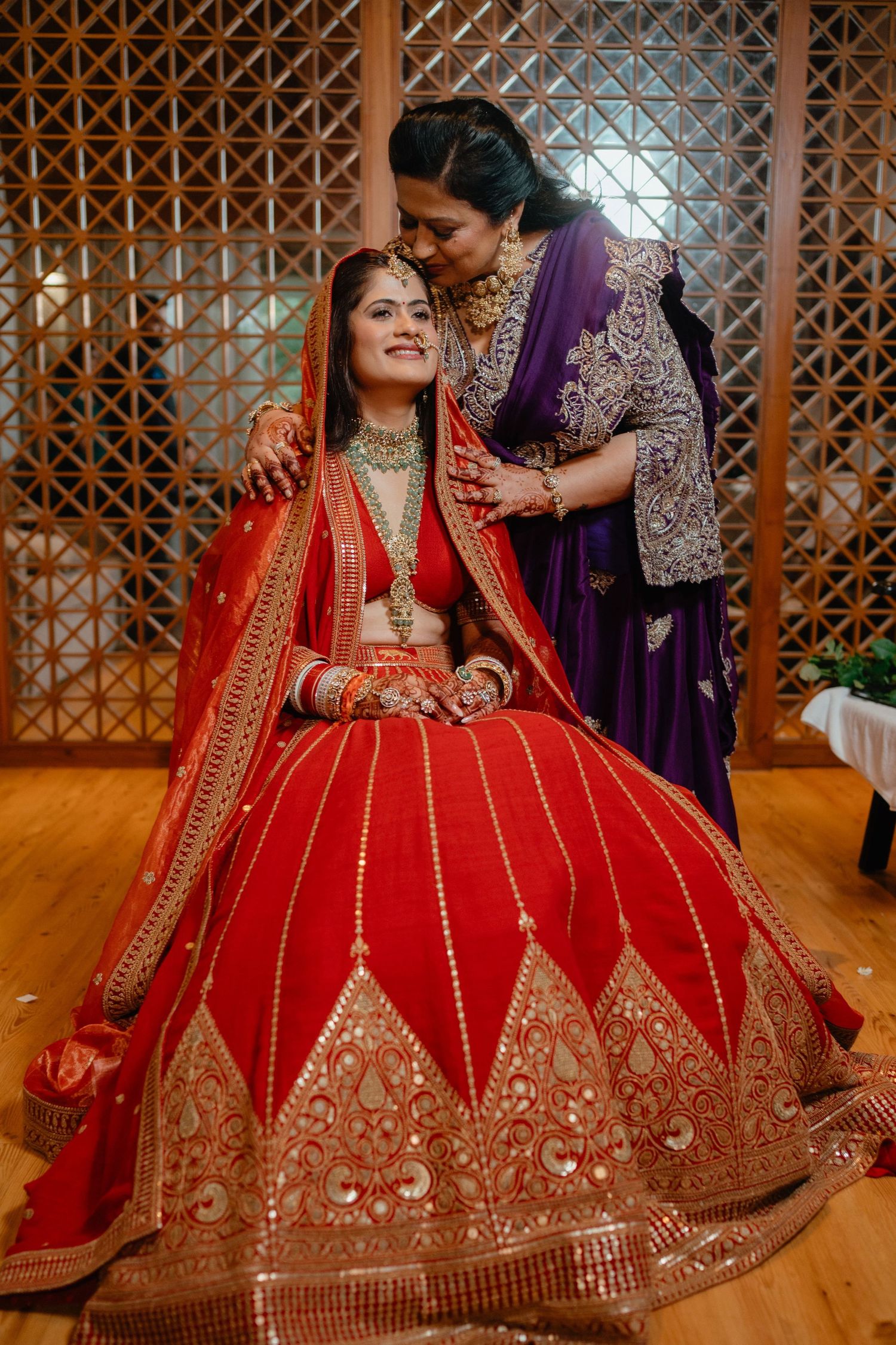 A bride in red lehenga with gold embroidery sits as an elder woman in purple saree blesses her at an Indian wedding.