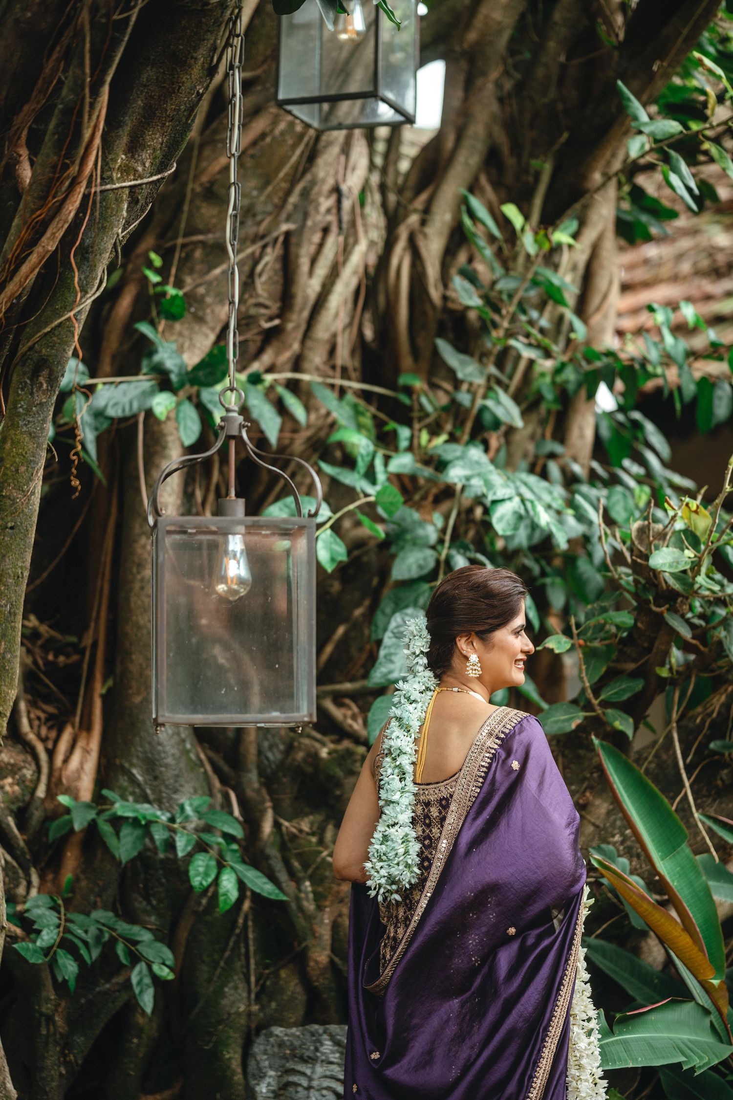 Woman in purple saree smiling near large banyan tree with hanging glass lanterns in lush garden setting.