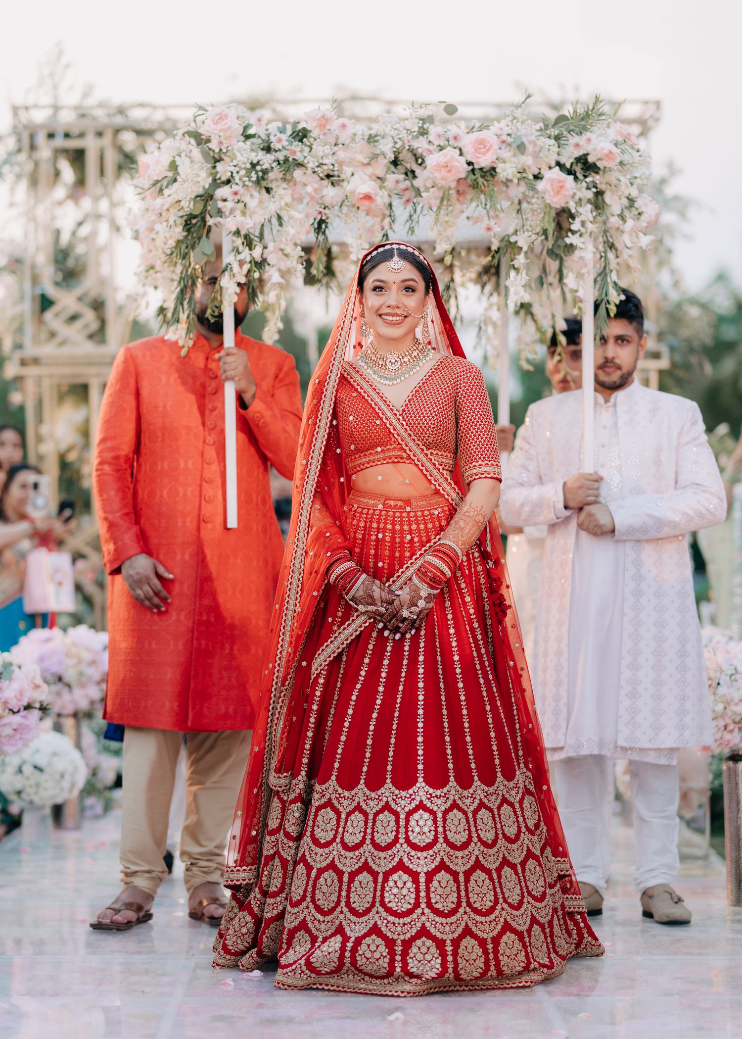 An Indian bride in a traditional red embroidered lehenga stands under a floral canopy at an outdoor wedding ceremony.