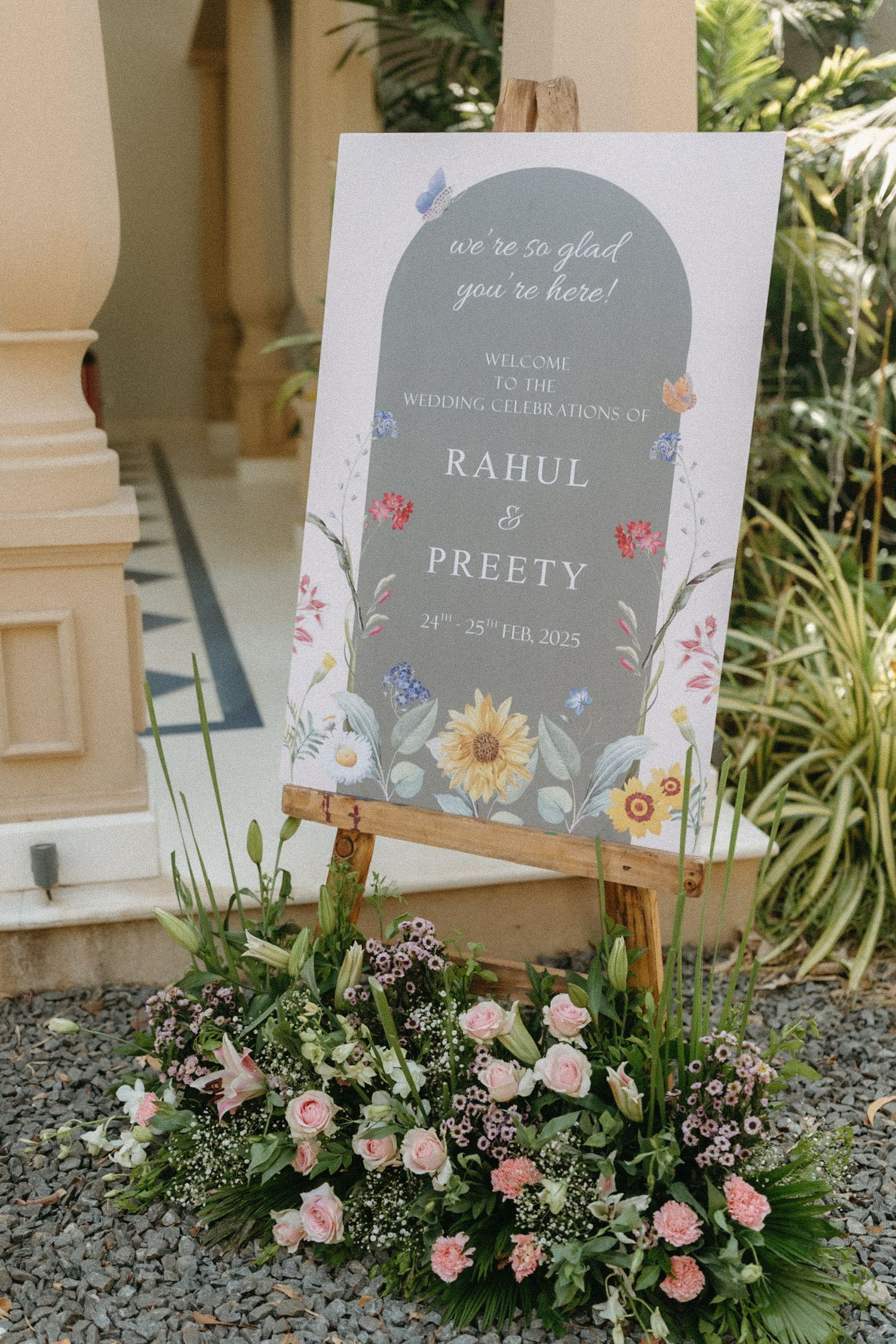 An elegant wedding welcome sign with floral design on a gray background stands on an easel surrounded by pink roses and greenery.