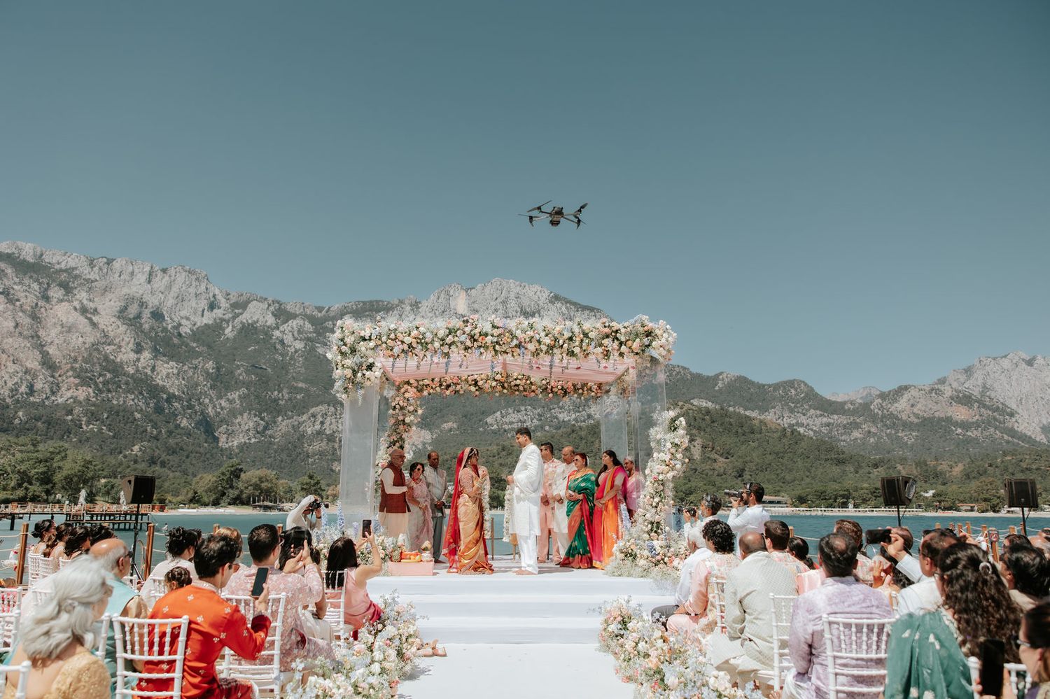 Outdoor Indian wedding ceremony with floral mandap, mountain lake backdrop, and drone flying overhead.