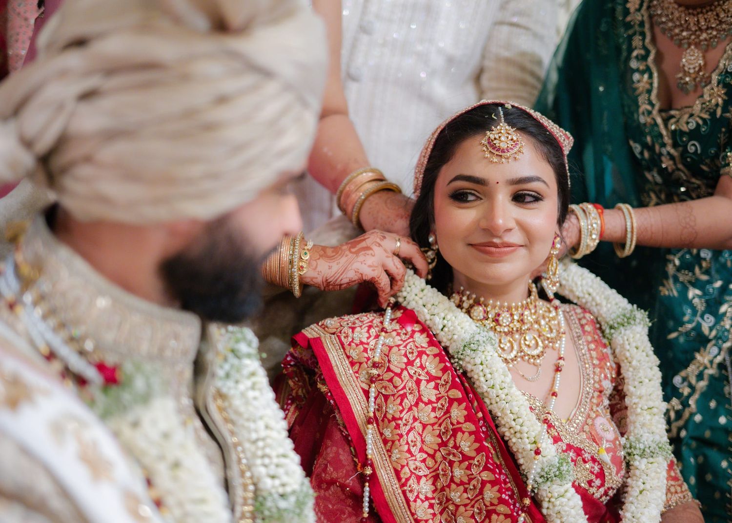 Bride in red lehenga with gold jewelry smiles during traditional Indian wedding ceremony.