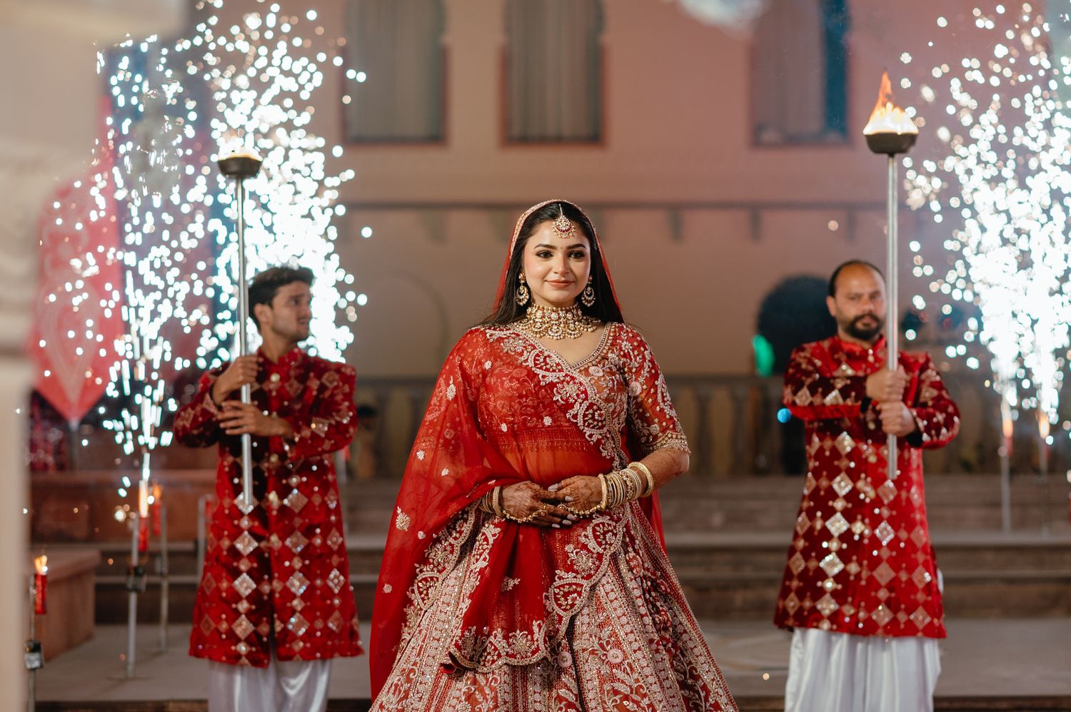 A bride in a red embroidered lehenga walks gracefully with sparklers and torch bearers at an Indian wedding ceremony.