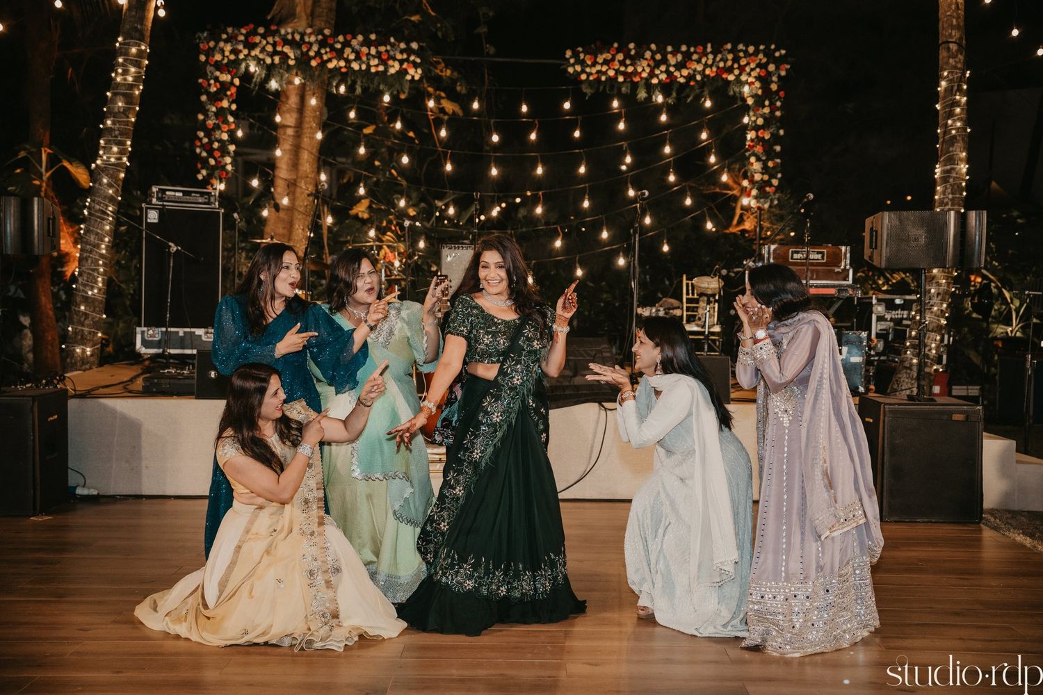 A group dances together in elegant formal attire under decorative string lights at an evening celebration.