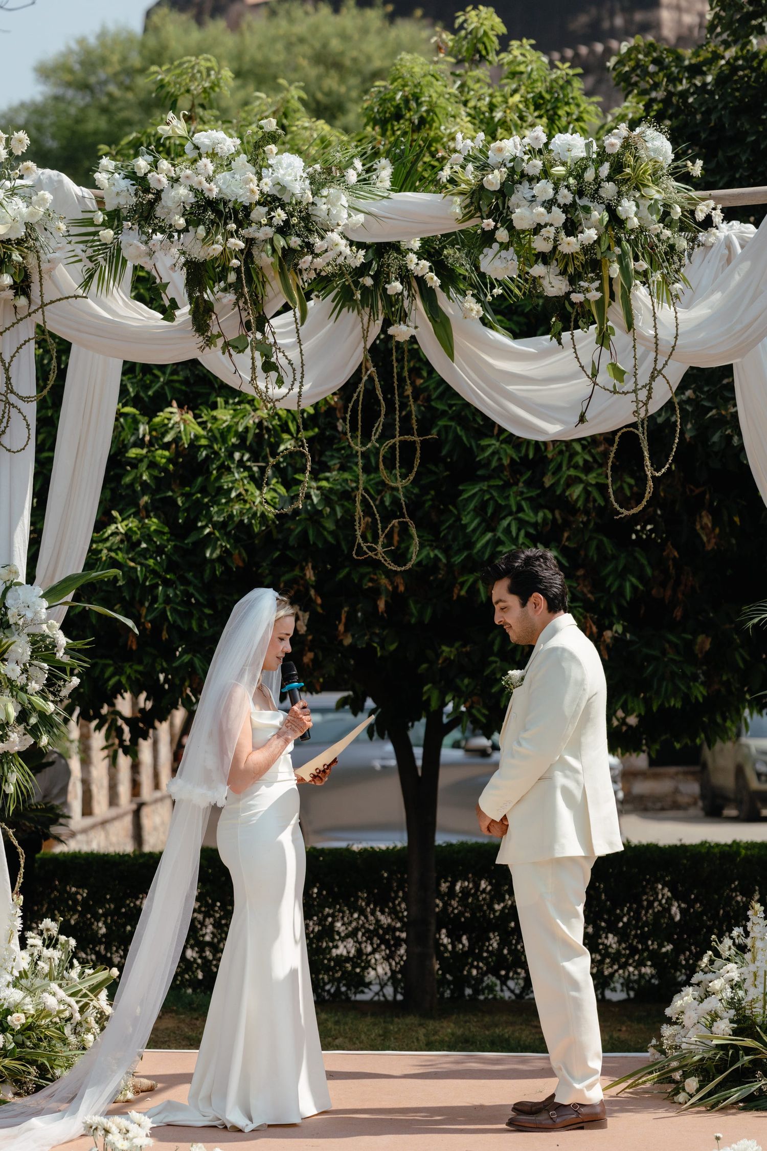 A wedding ceremony under an elegant white floral arch with flowing fabric draping and greenery in an outdoor garden setting.