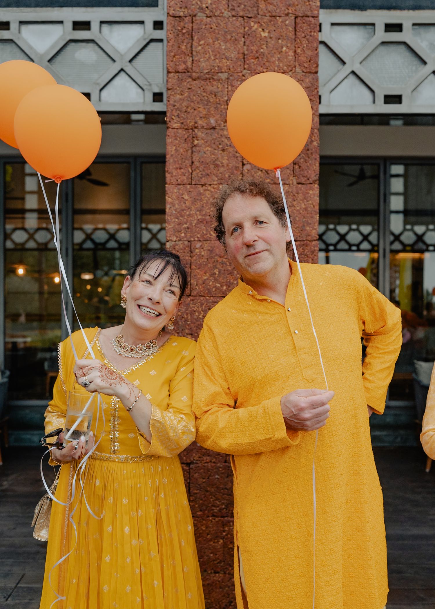 Couple in matching yellow outfits poses with orange balloons against a brick and metal backdrop.