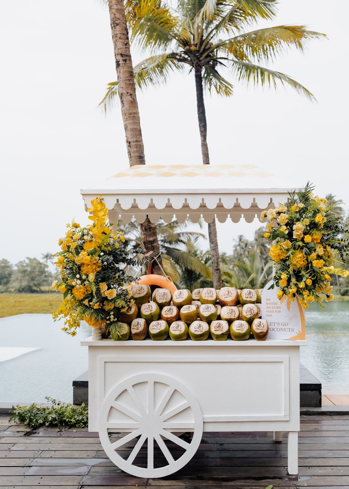 White decorative cart with fresh coconuts and yellow flower arrangements under palm trees.