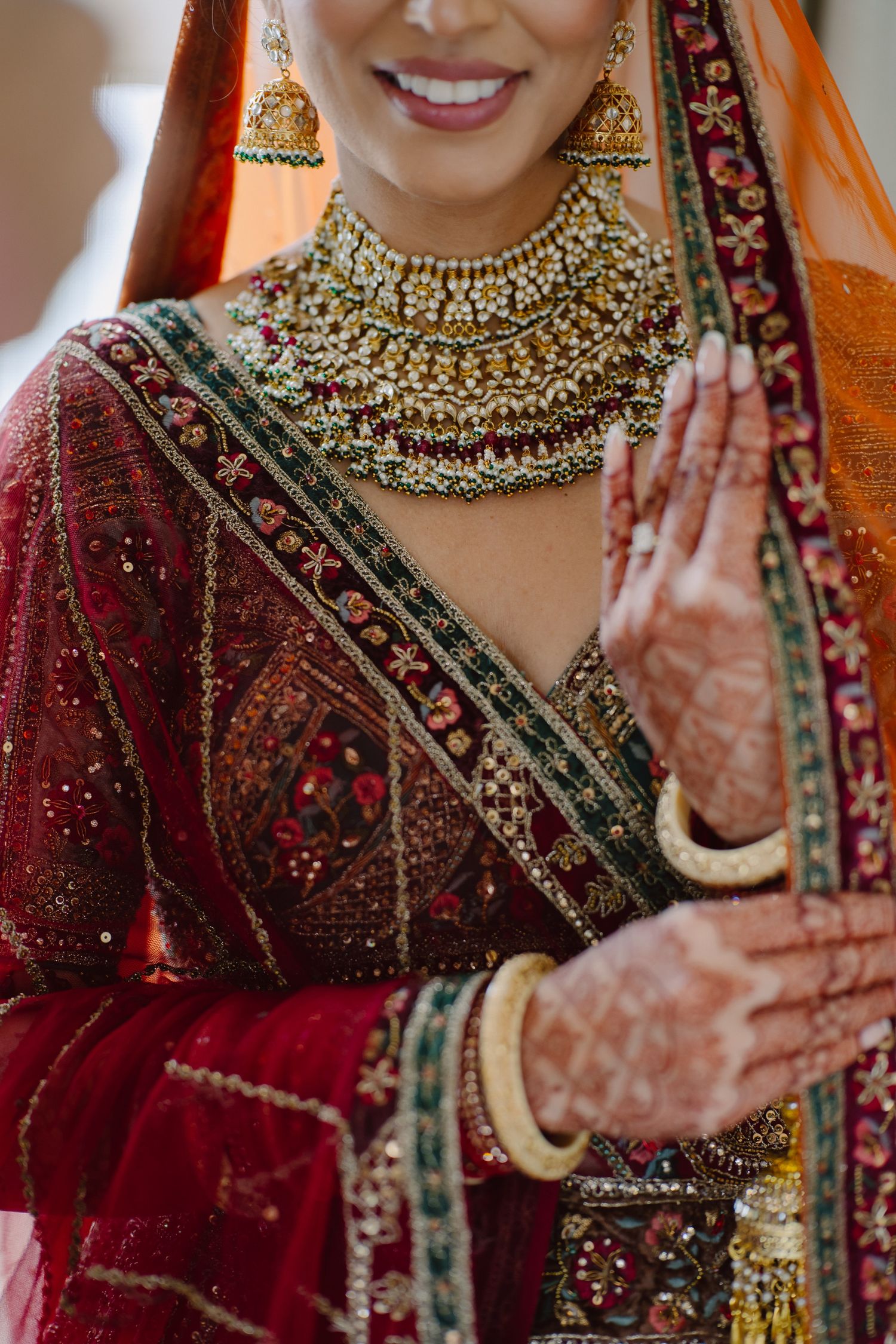 Indian bride in red lehenga with gold kundan jewelry, mehndi hands, and orange dupatta smiling at wedding.
