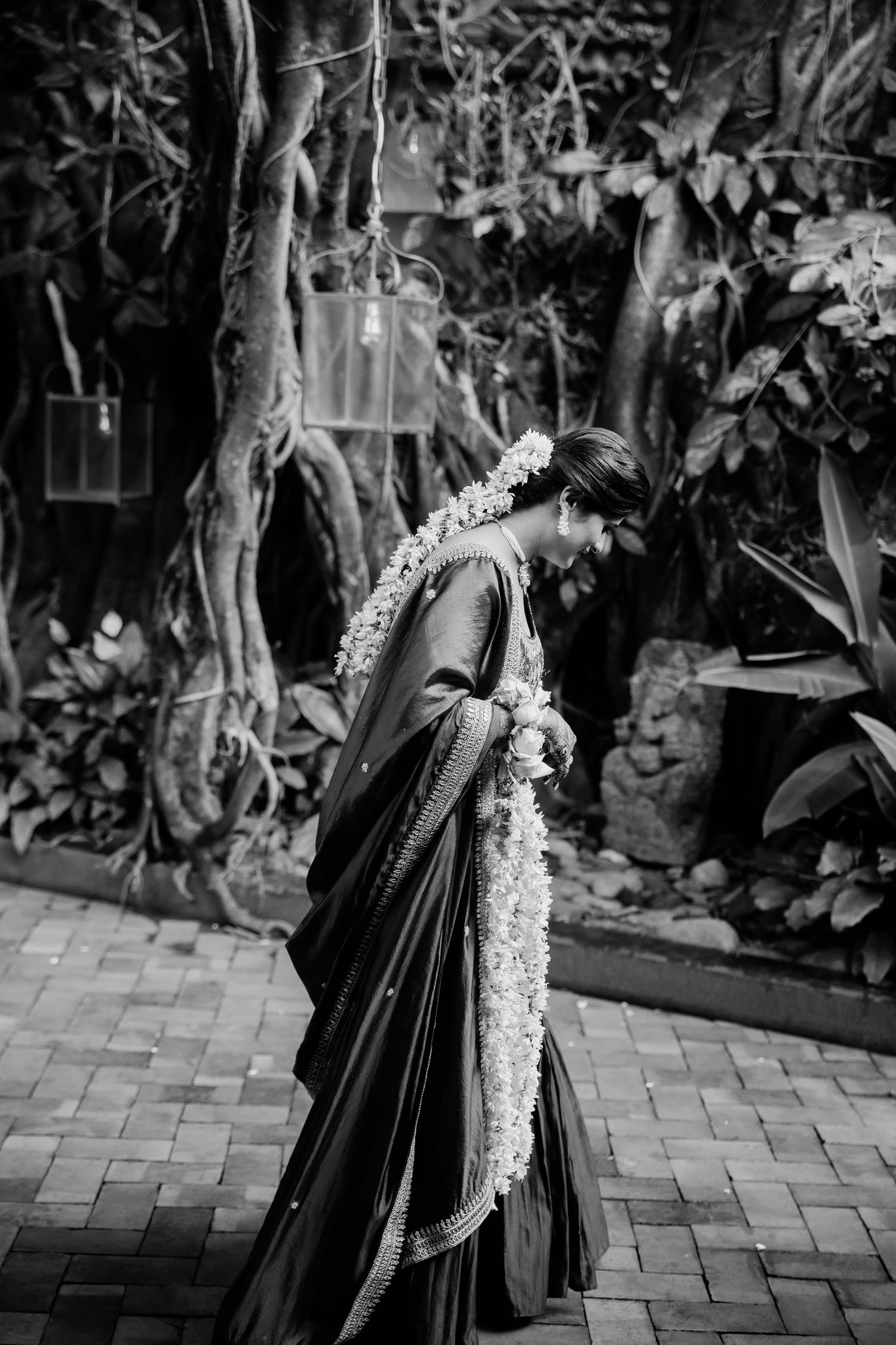 Black and white photo of a woman in a saree with floral garland, standing in a lush garden setting.