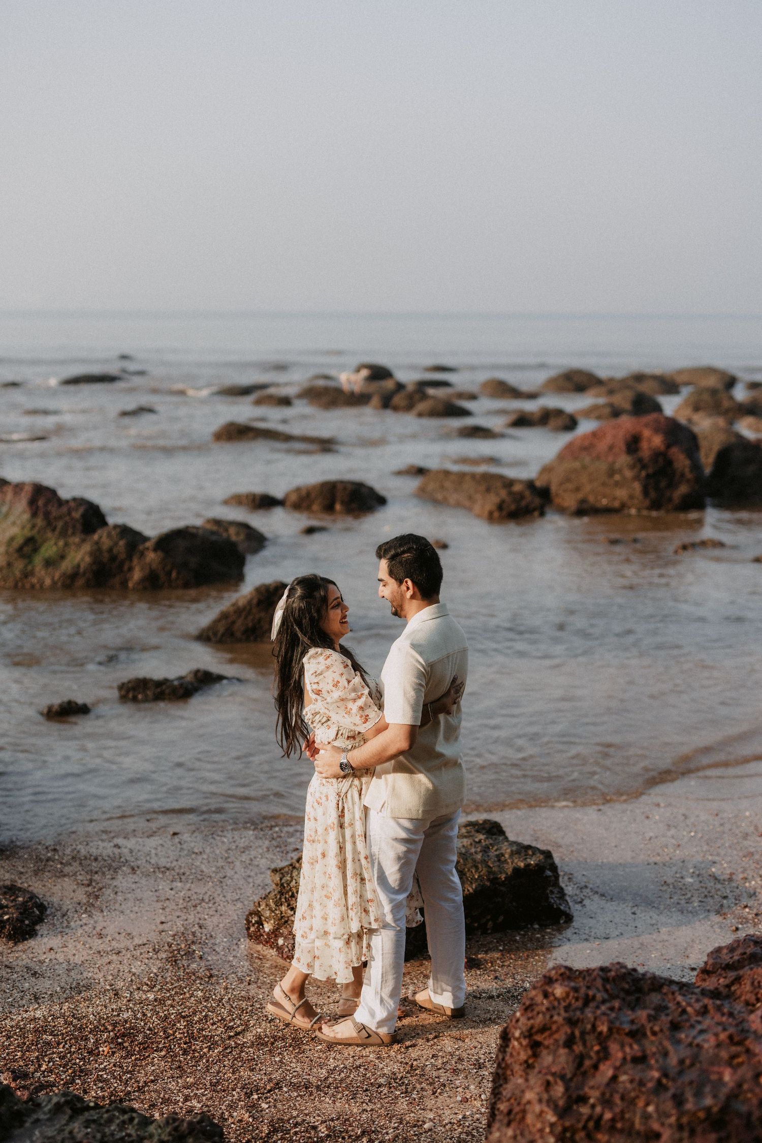 A couple embraces on a rocky beach at sunset with waves and boulders in the misty background.