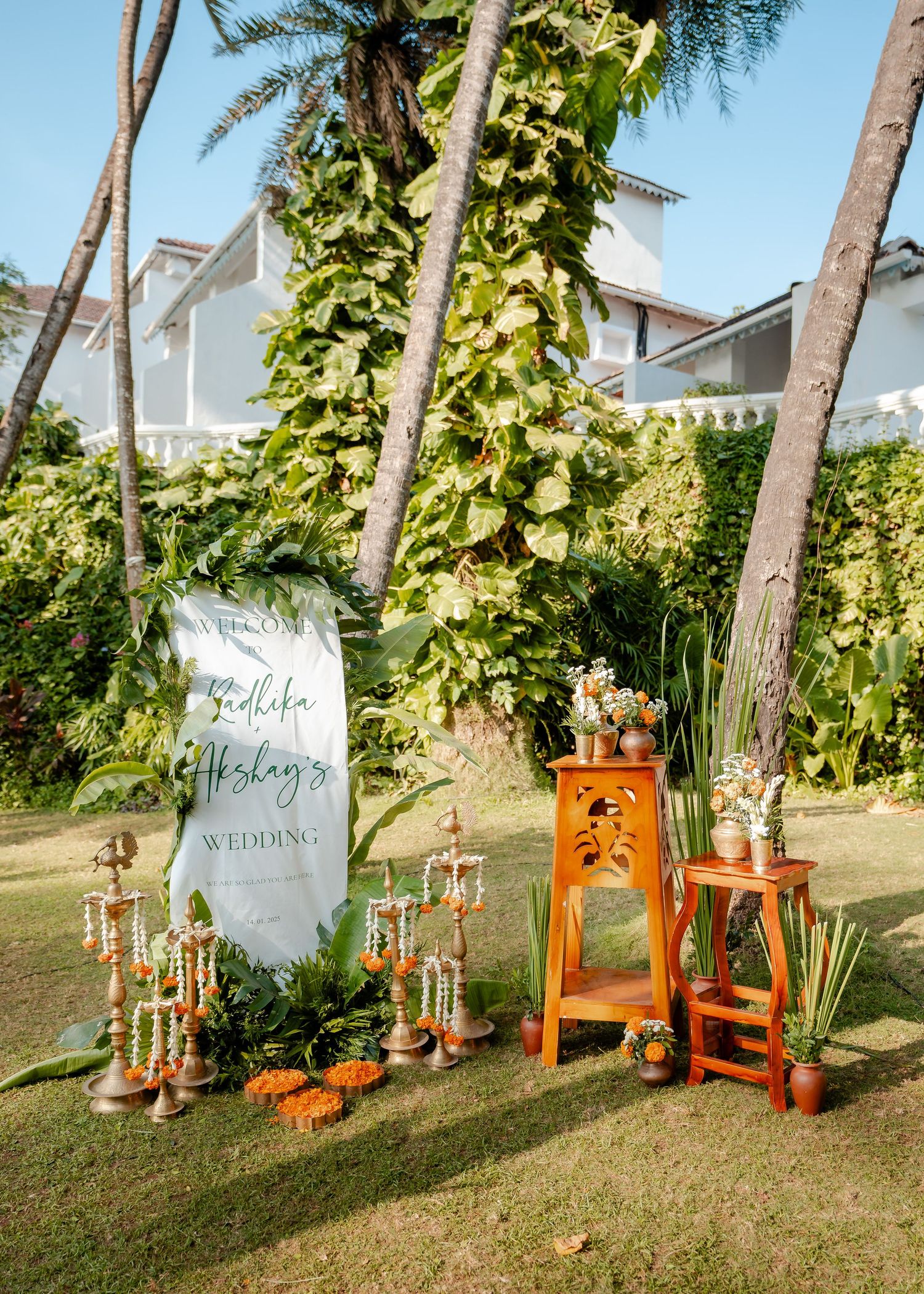 Outdoor Indian wedding welcome sign with marigold garlands, wooden stands, and tropical palm trees in a lush garden setting.