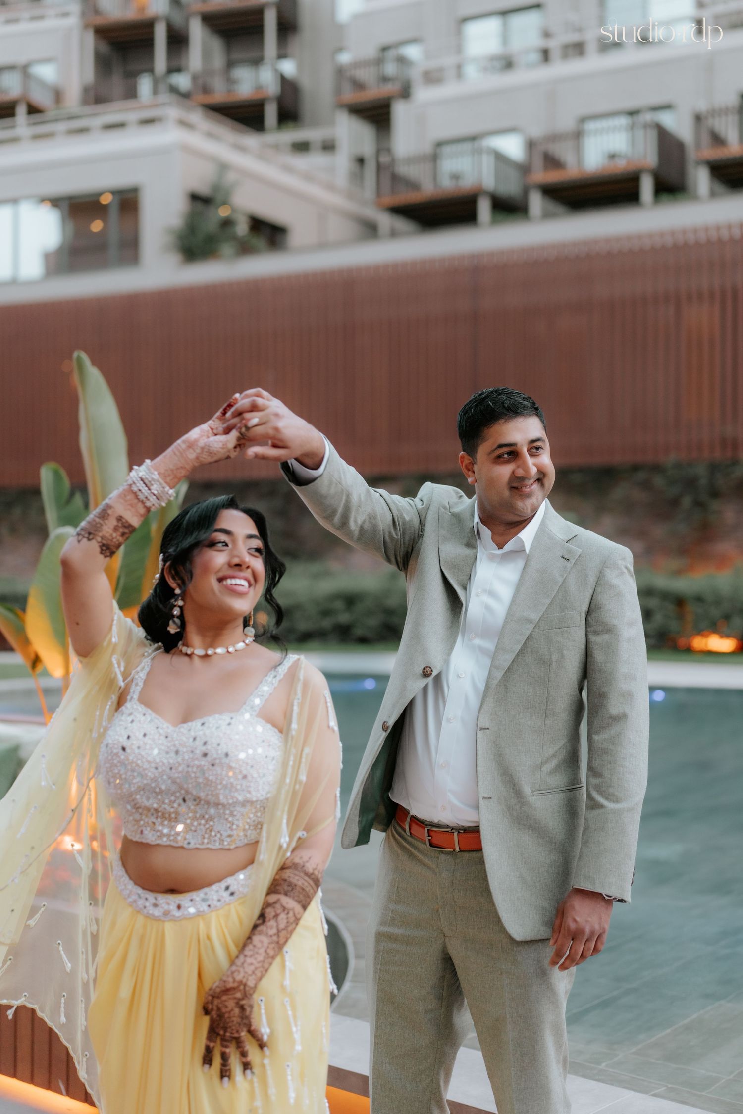 Couple in Indian wedding attire dancing outdoors, bride in yellow lehenga with mehndi, groom in grey suit by poolside.