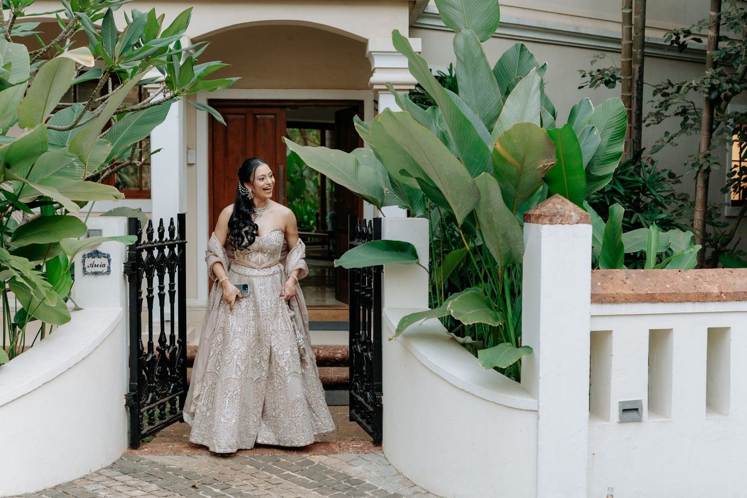 Woman in elegant lehenga standing at ornate gate entrance with tropical plants and white colonial architecture.