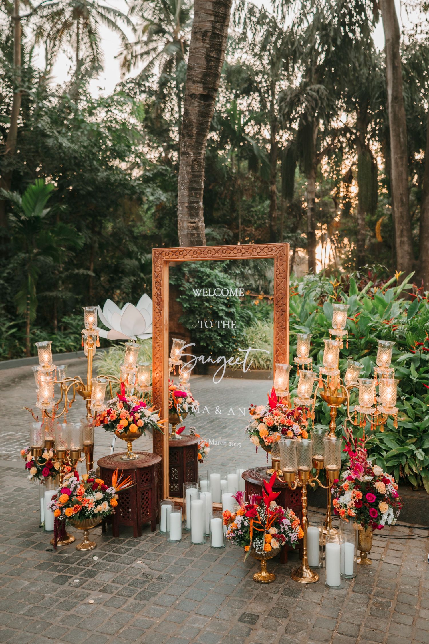 Elegant outdoor wedding display with wooden frames, white candles, and vibrant coral flower arrangements in a tropical garden setting.