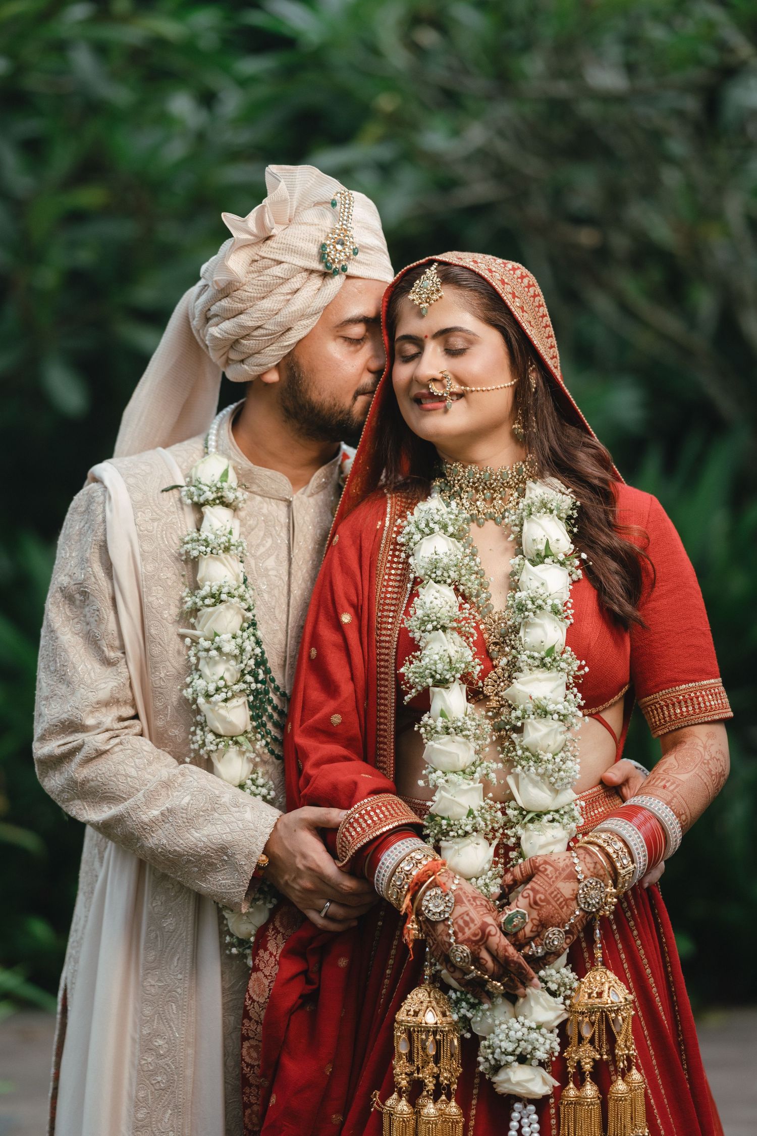 Indian groom in cream sherwani kisses bride in red lehenga wearing white floral garlands at wedding.