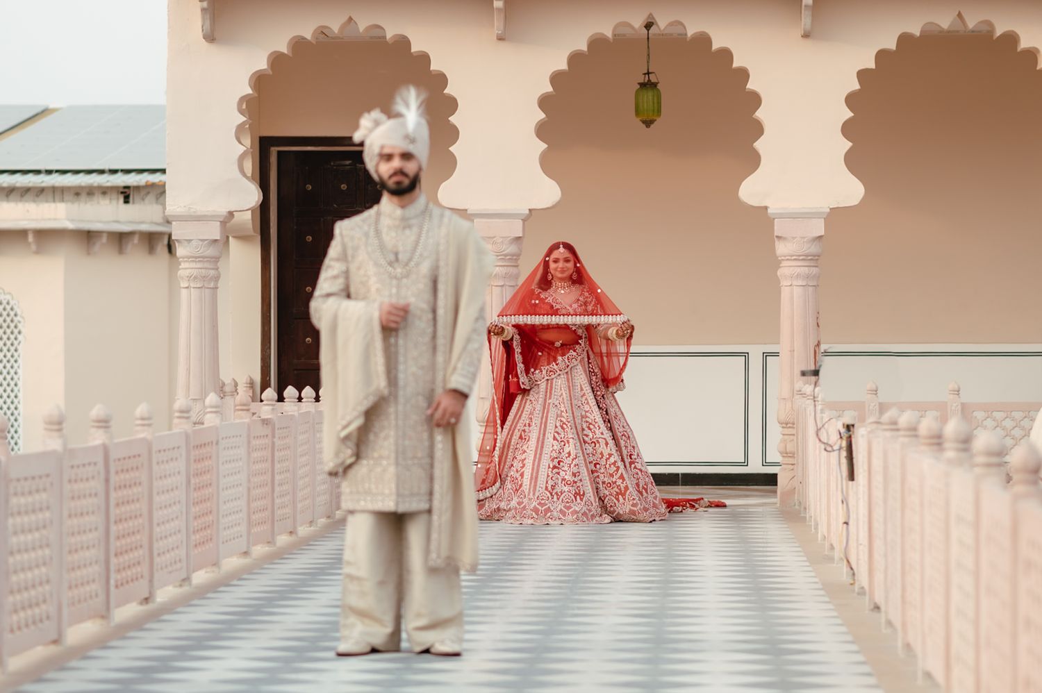 Indian groom in white sherwani and bride in red lehenga pose at ornate palace venue for traditional wedding.