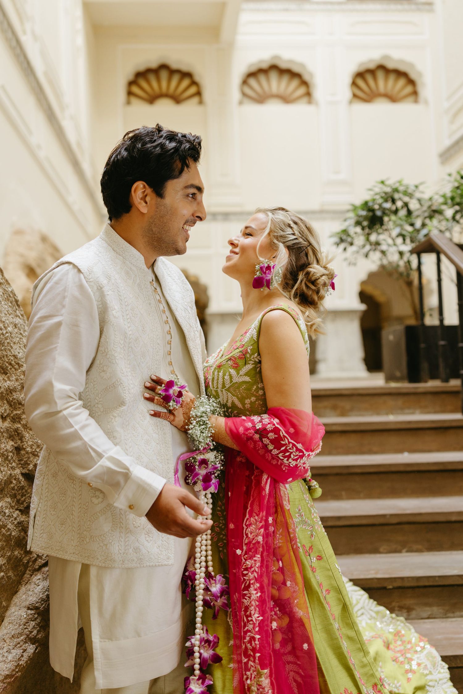 A couple in white and green traditional attire pose on palace steps with pink details.