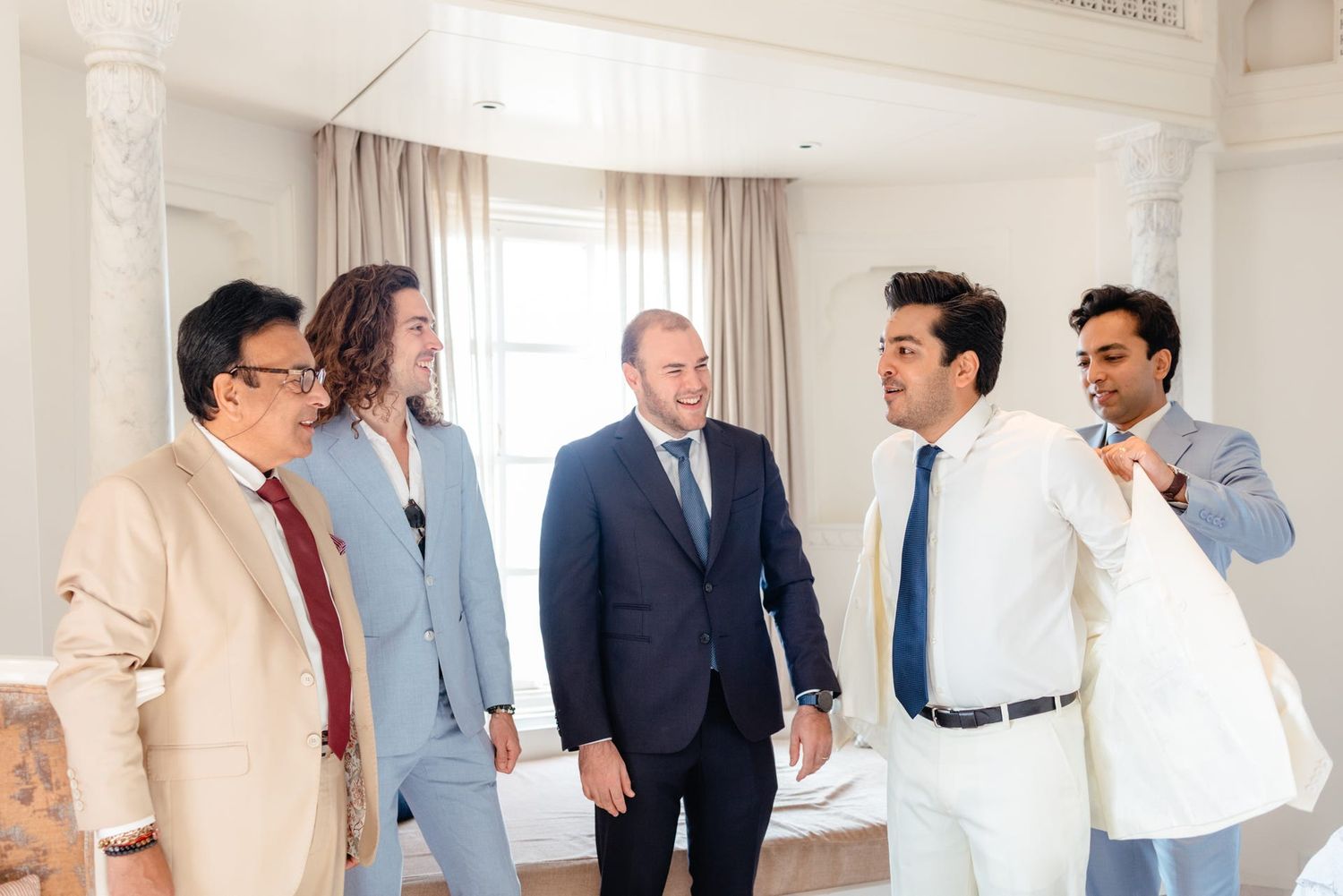 Wedding party members in light colored suits gathered in an elegant white room.
