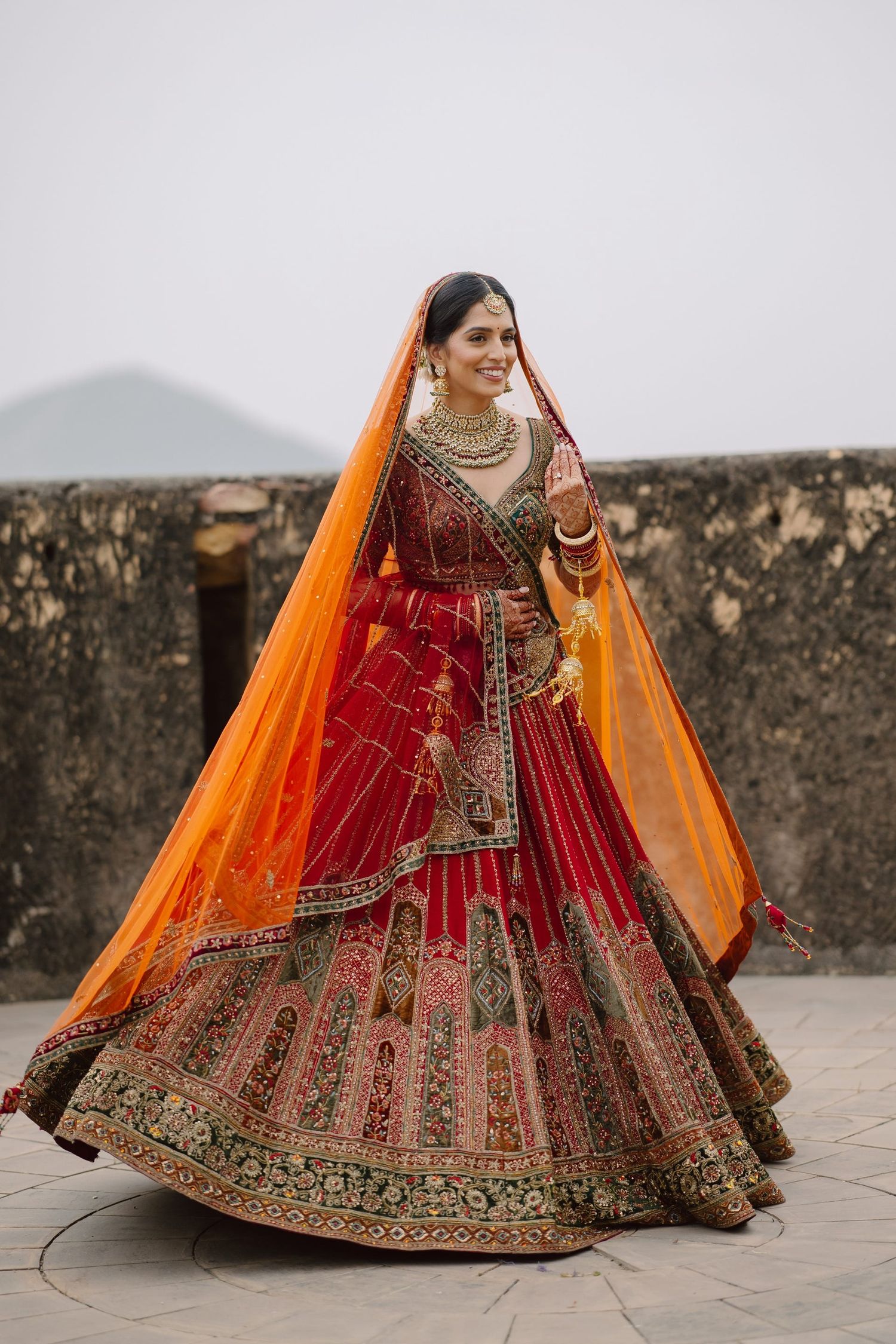 Ornate red and orange bridal lehenga with intricate embroidery photographed against stone wall.