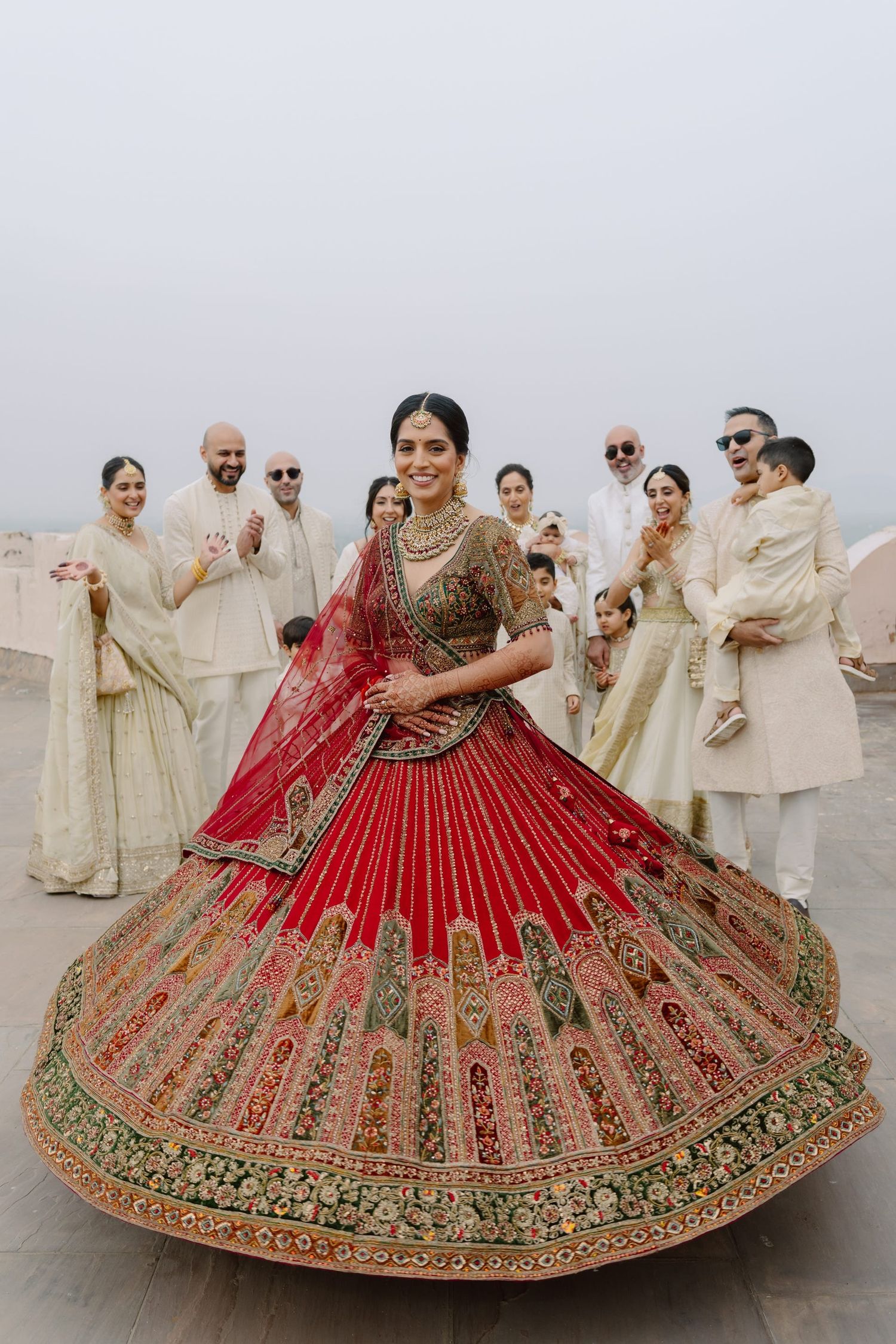 A bride in a red embroidered lehenga twirls while surrounded by guests in cream outfits.