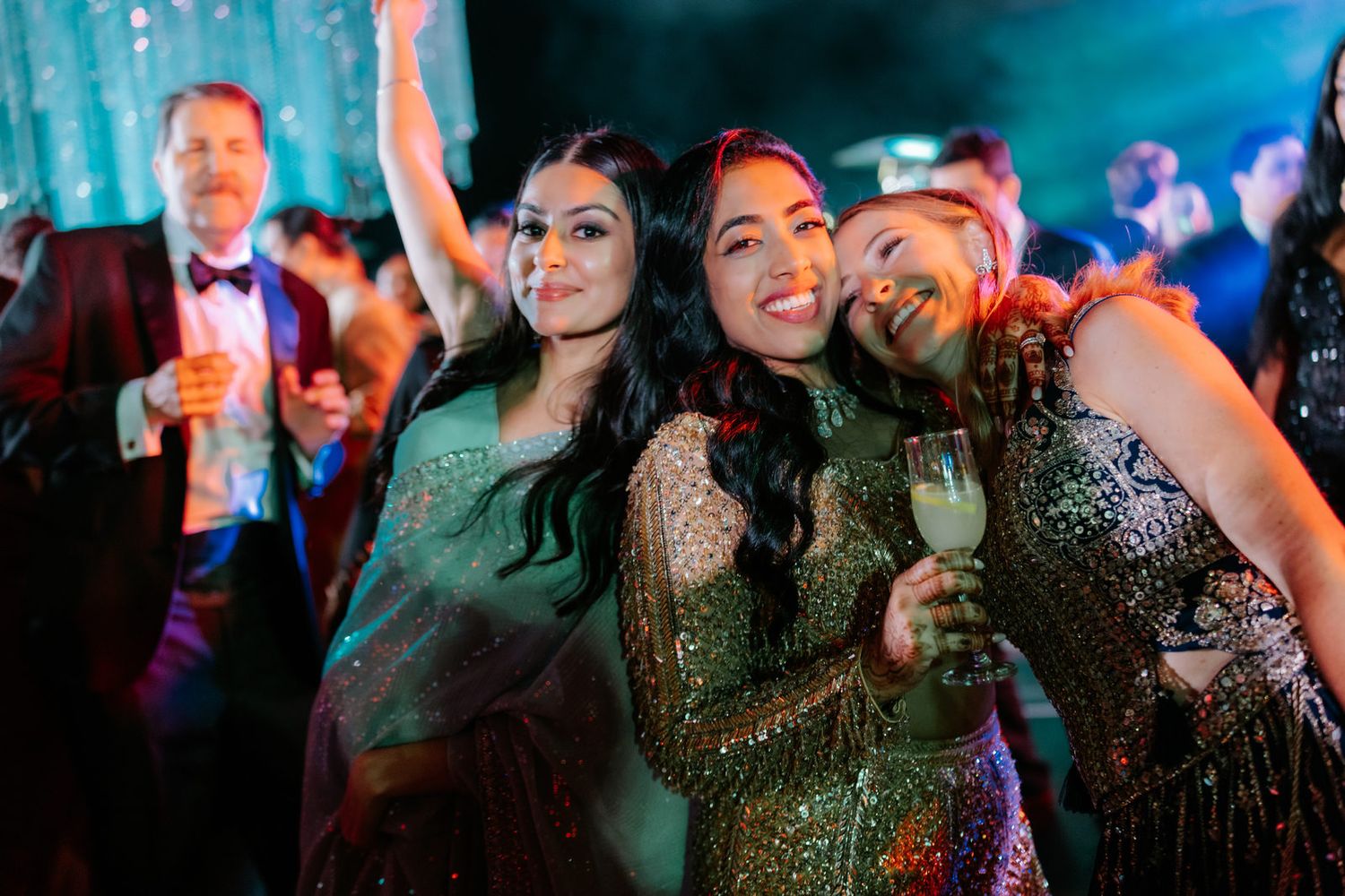 Three women in sequin dresses smile and pose together at a glamorous party event with colorful lighting.