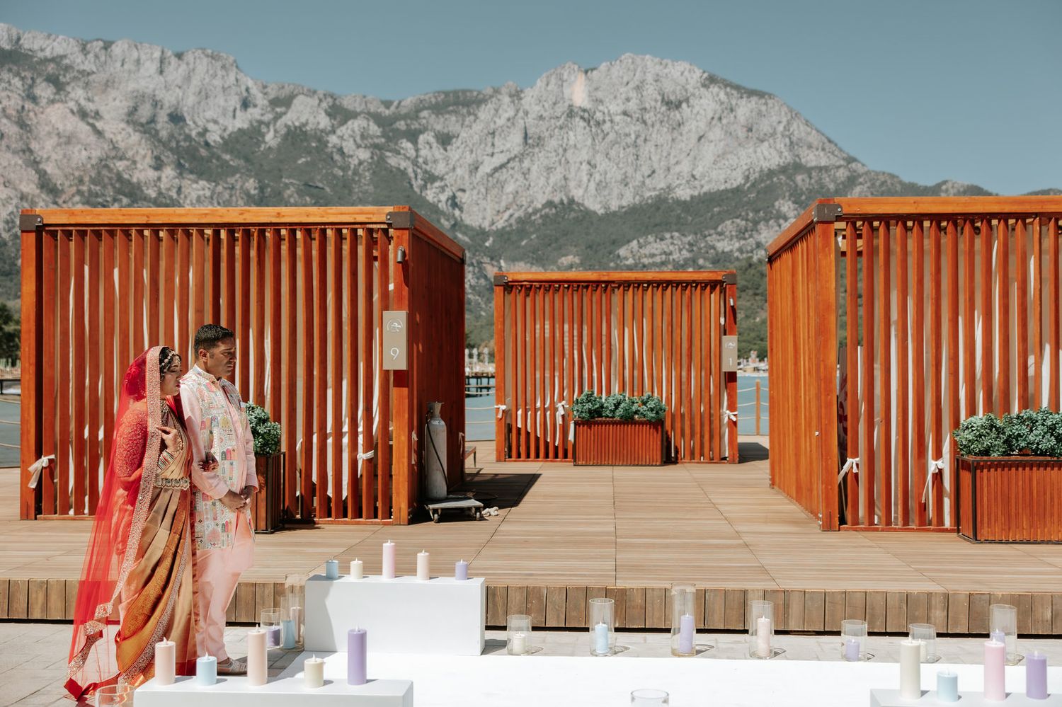 Bride and groom in traditional Indian attire share a moment by wooden pavilions with mountain backdrop.