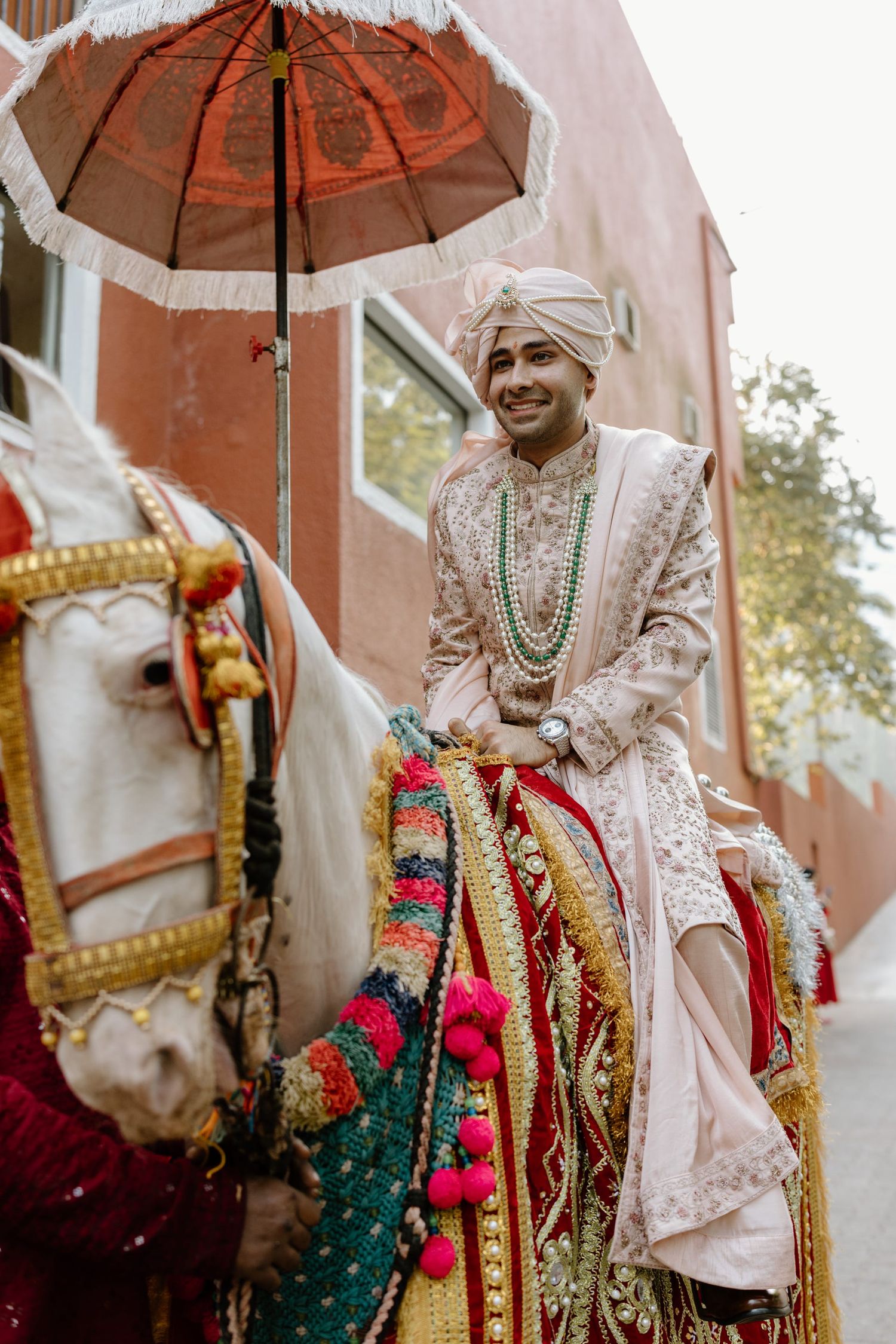 Traditional Indian groom wearing ornate white sherwani and riding a decorated white horse under a red umbrella.