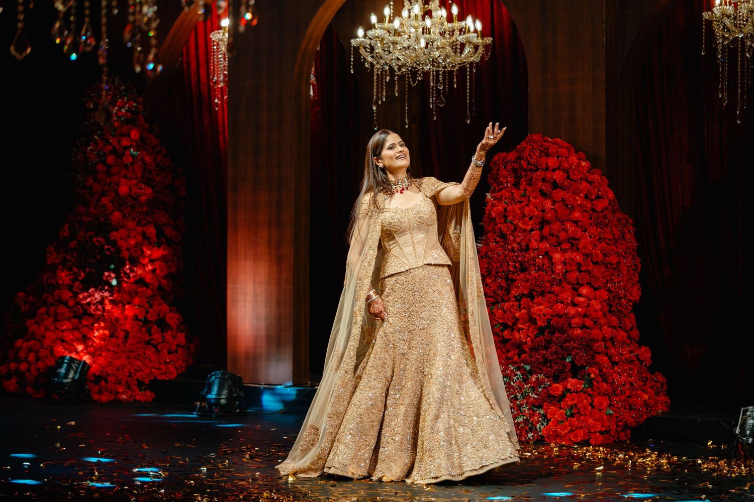 Woman in gold lehenga poses elegantly on stage with red floral arrangements and chandelier backdrop.