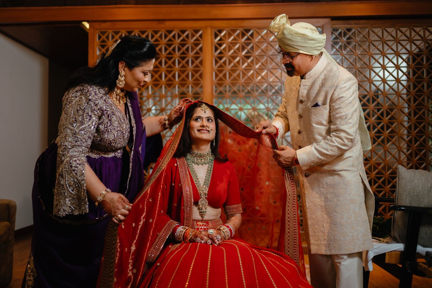 Indian bride in red lehenga being helped with dupatta by family members during traditional wedding preparation ceremony.